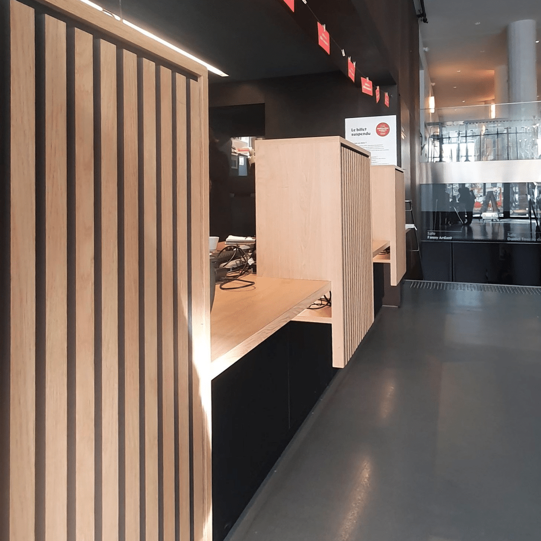 Comptoir d'accueil moderne en bois clair avec lamelles verticales, signalisation "Le billet suspendu" et vue sur un hall vitré.