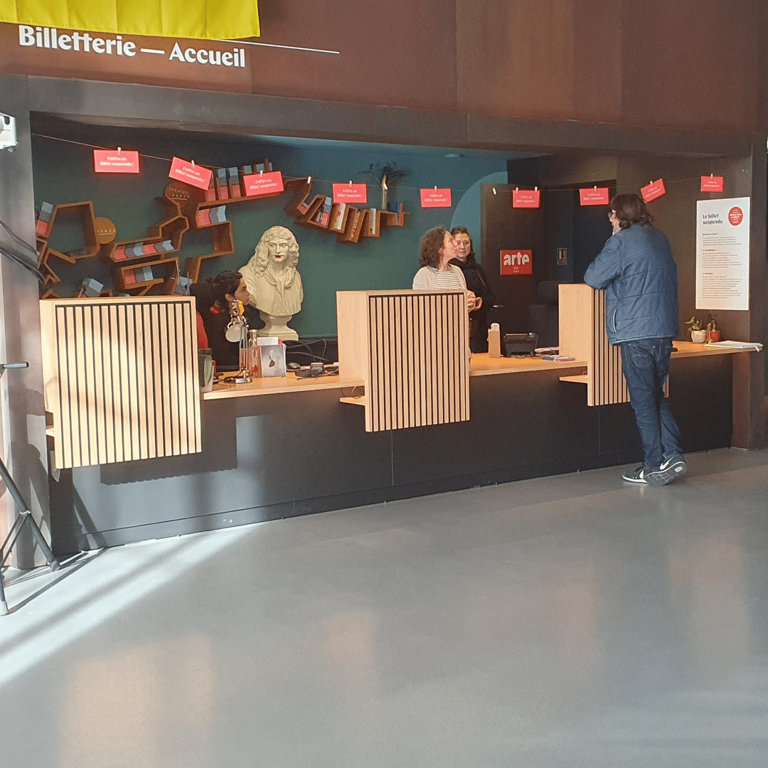 Guichet de billetterie-accueil avec une inscription jaune et un buste sculpté, des employés servant un client. Panneau 'Le billet suspendu'.