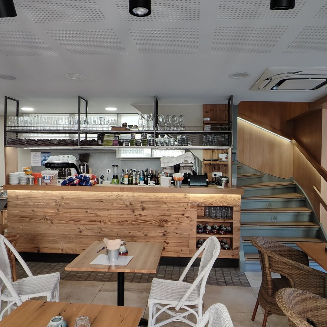 Intérieur de café moderne avec comptoir en bois clair, machines à café, étagères chargées de verres et d'alcool, et escalier adjacent en bois clair et marches bleues.