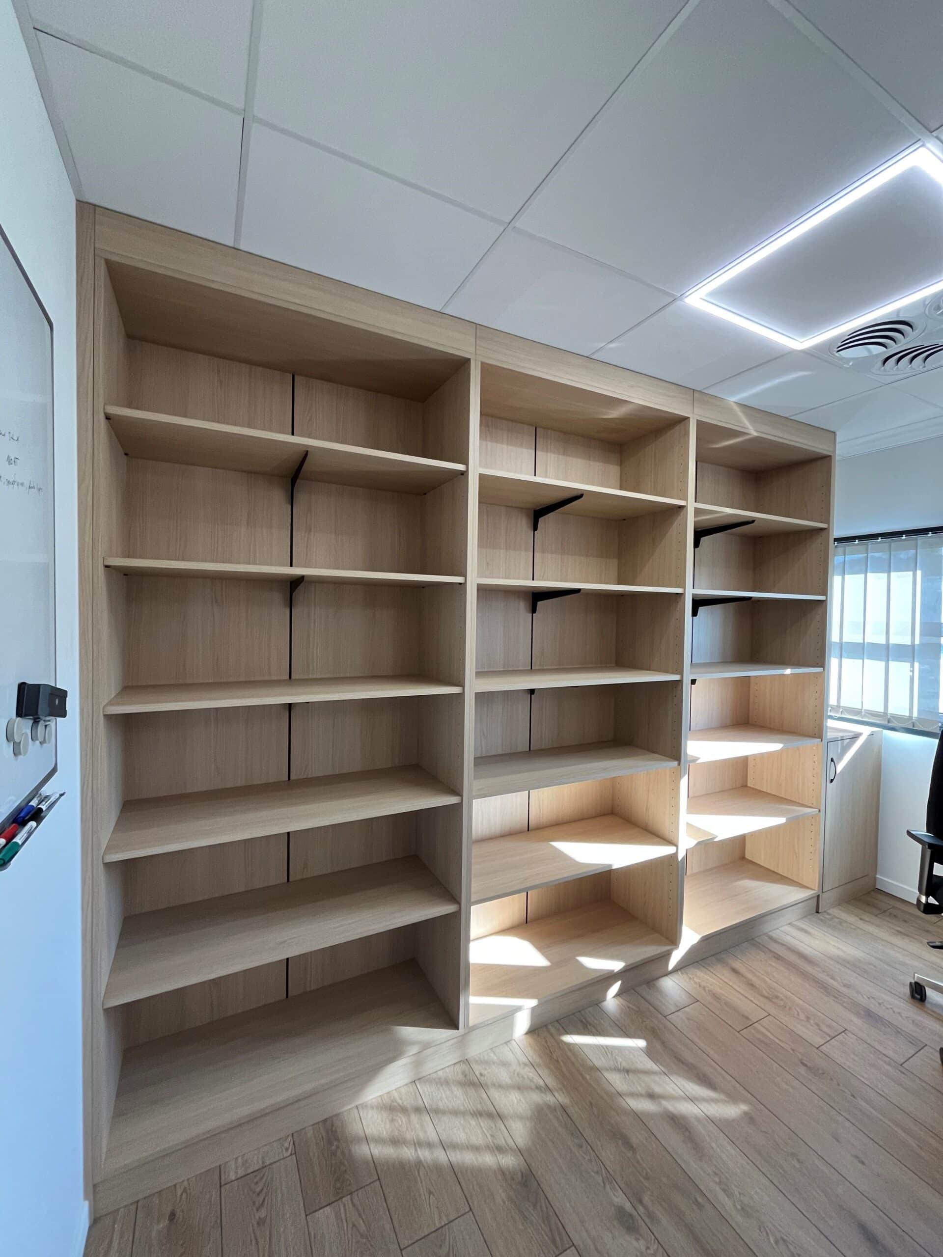 Bibliothèque murale en bois clair vide dans un bureau moderne avec éclairage naturel.