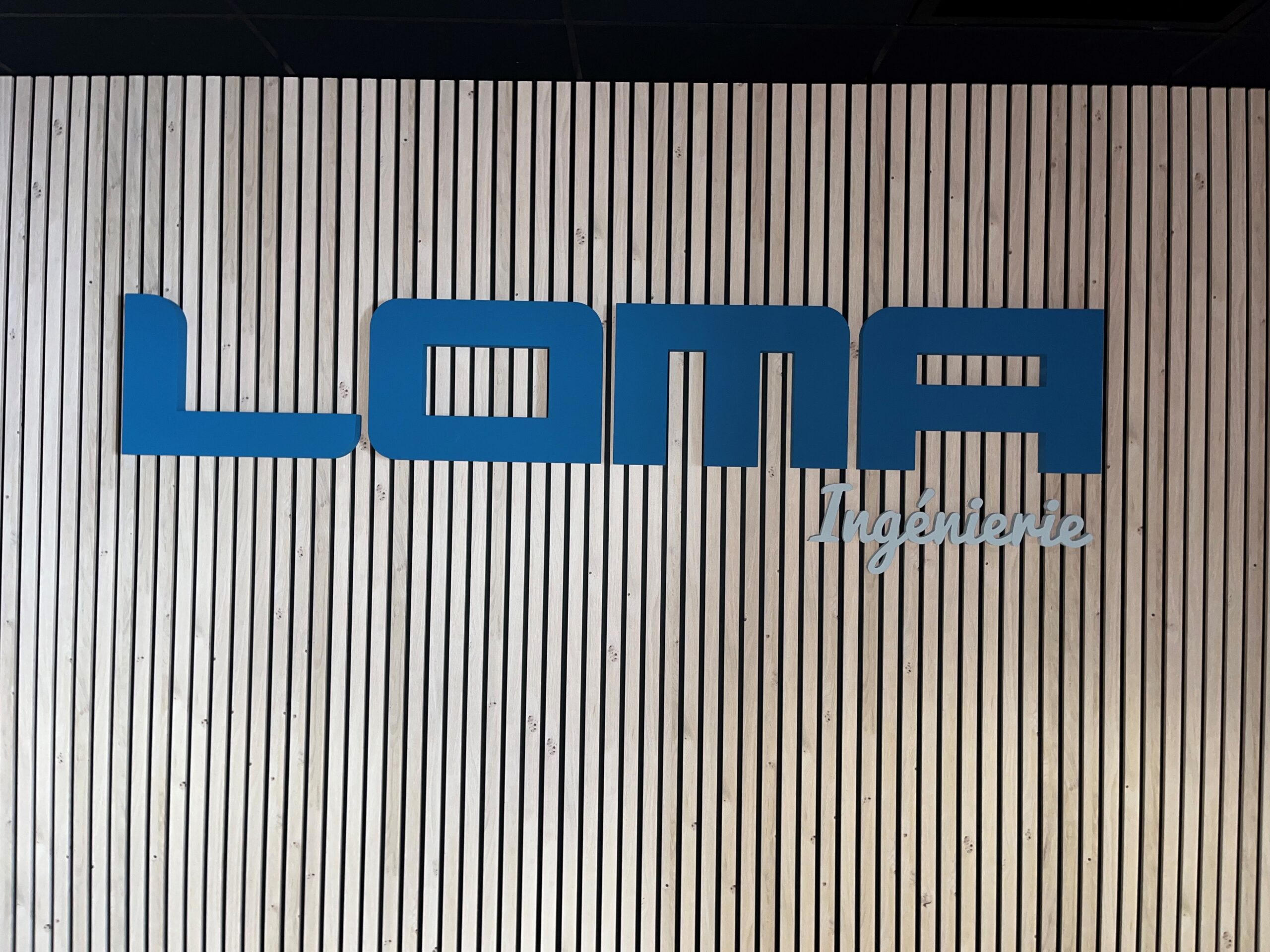 Logo bleu LOMA Ingénierie fixé sur un mur décoratif en tasseaux de bois clair.