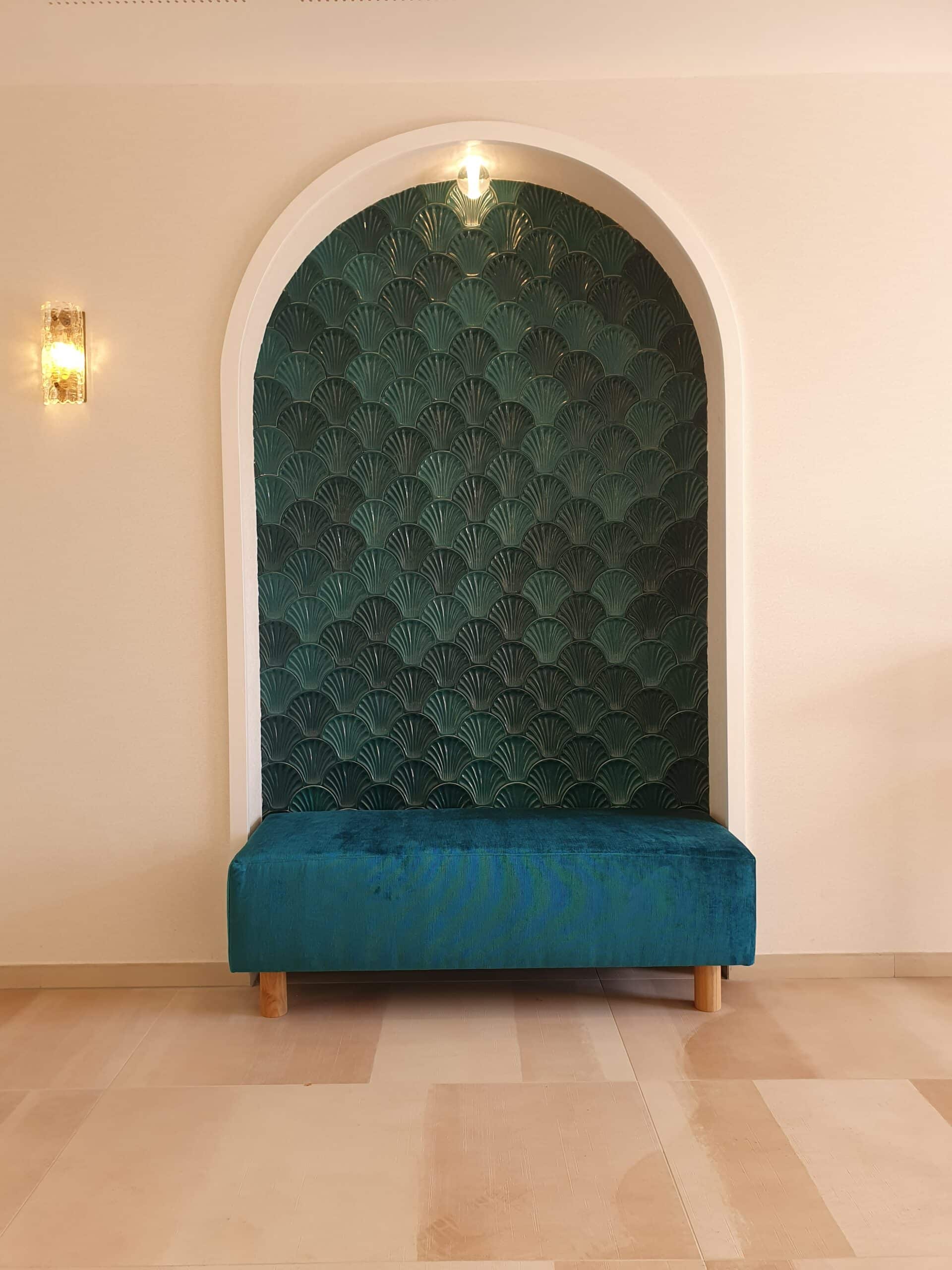 Banquette velours bleu devant une niche en carrelage écailles vert émeraude sous une arche éclairée.