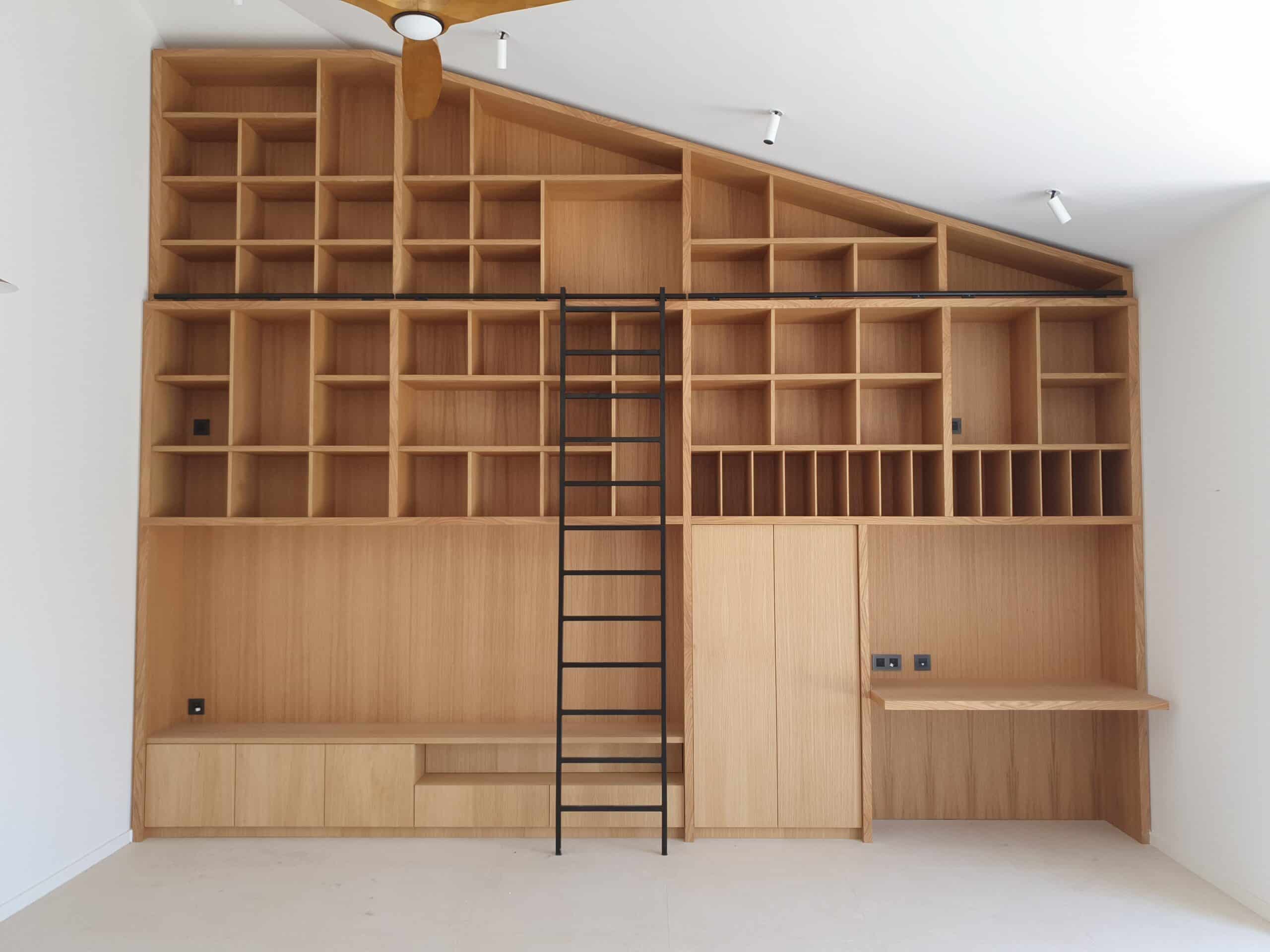 Bibliothèque sur mesure en bois clair, échelle noire et bureau intégré sous un plafond mansardé.