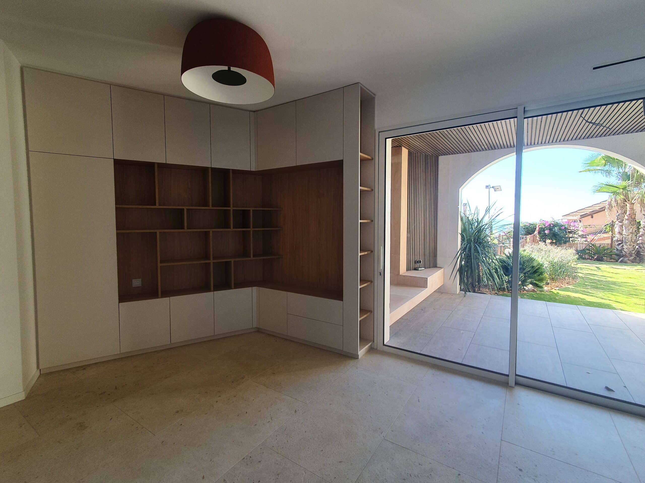 Salon moderne avec bibliothèque sur mesure, suspension rouge et grande baie vitrée ouvrant sur un jardin ensoleillé.