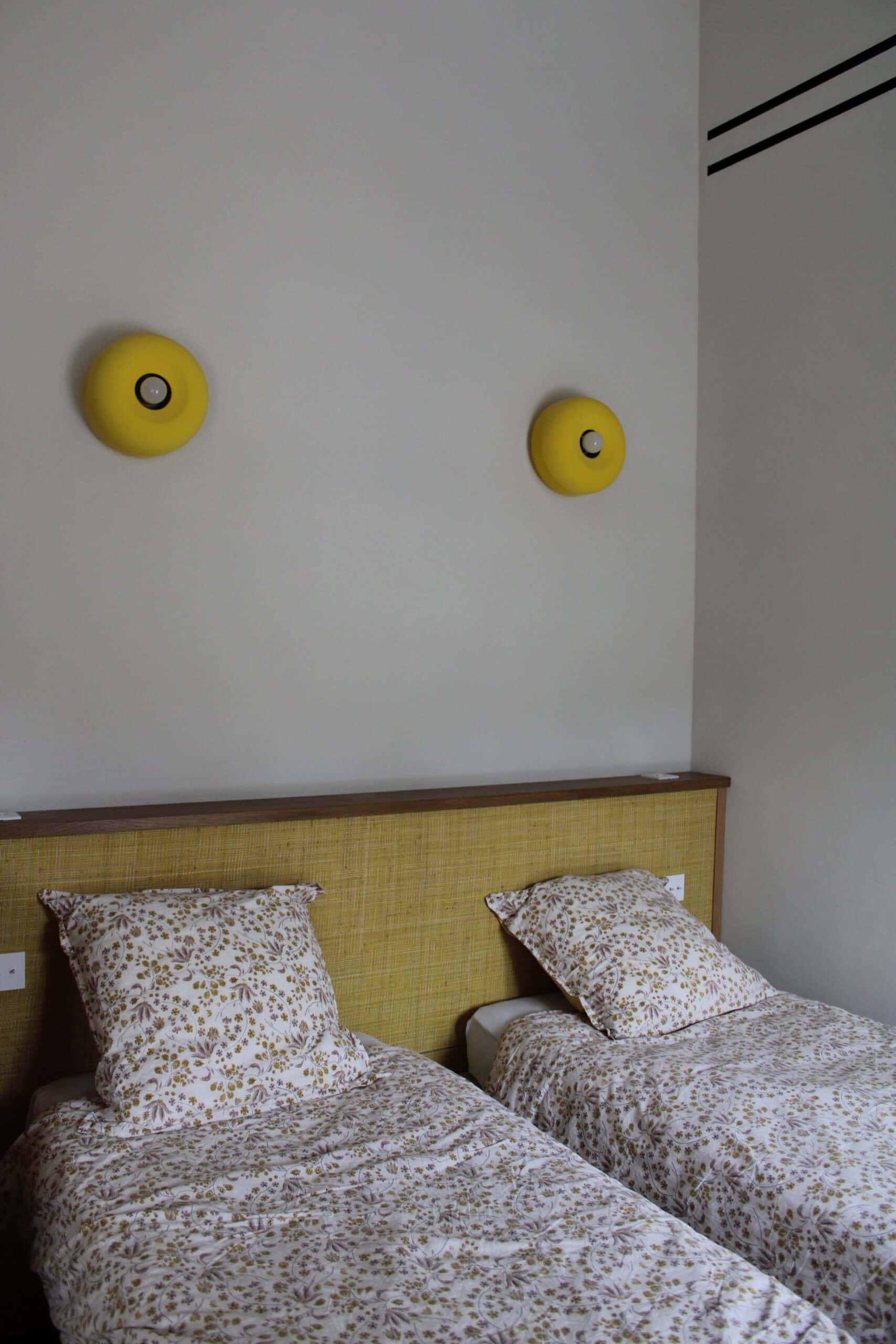 Deux lits jumeaux, couettes fleuries, tête de lit jaune et appliques murales assorties sur mur blanc.