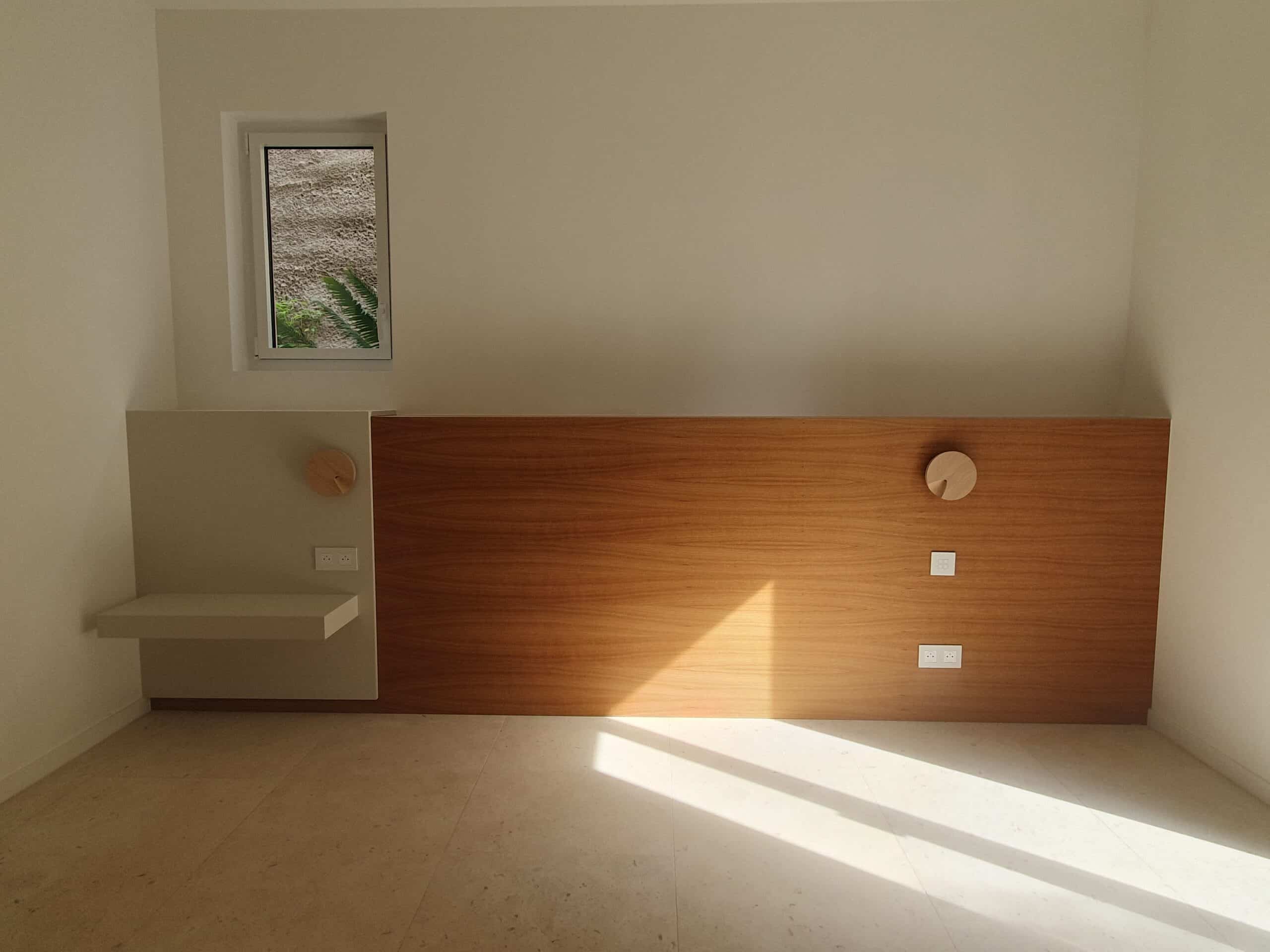Tête de lit en bois massif, mur blanc épuré, étagère intégrée et lumière naturelle rasante au sol.