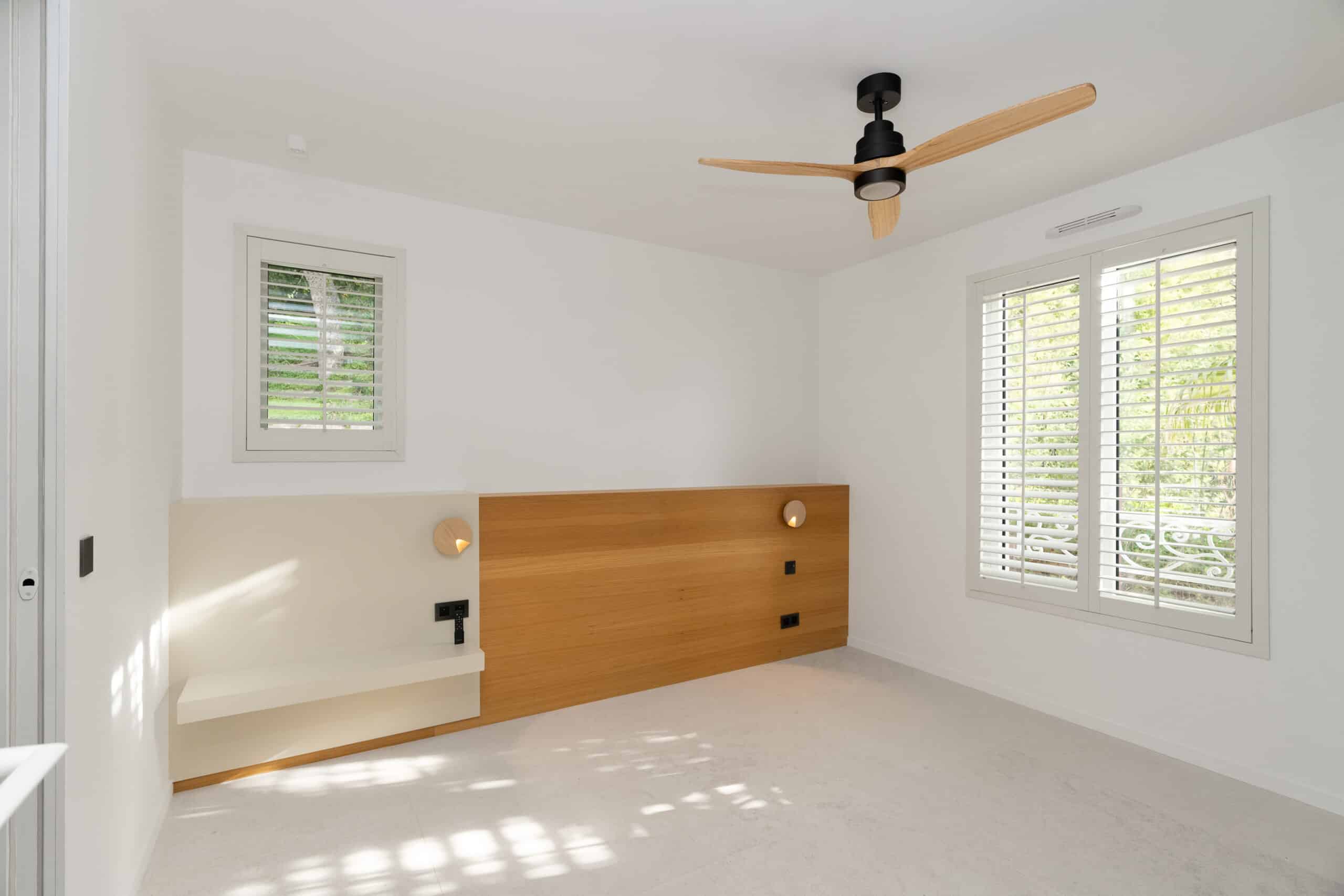 Chambre moderne blanche avec tête de lit en bois et ventilateur de plafond design.