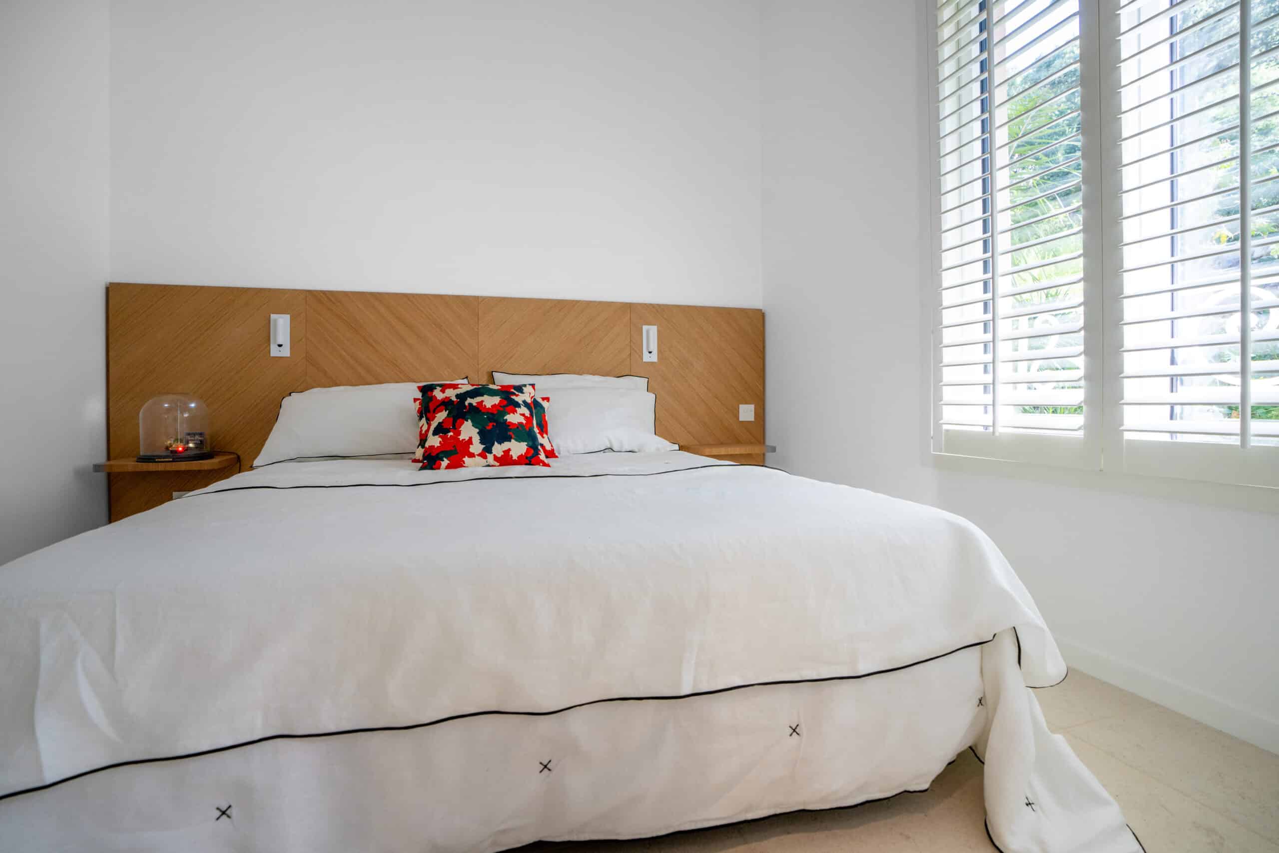 Chambre moderne avec lit blanc, tête de lit en bois, coussin coloré et fenêtre à persiennes blanches.