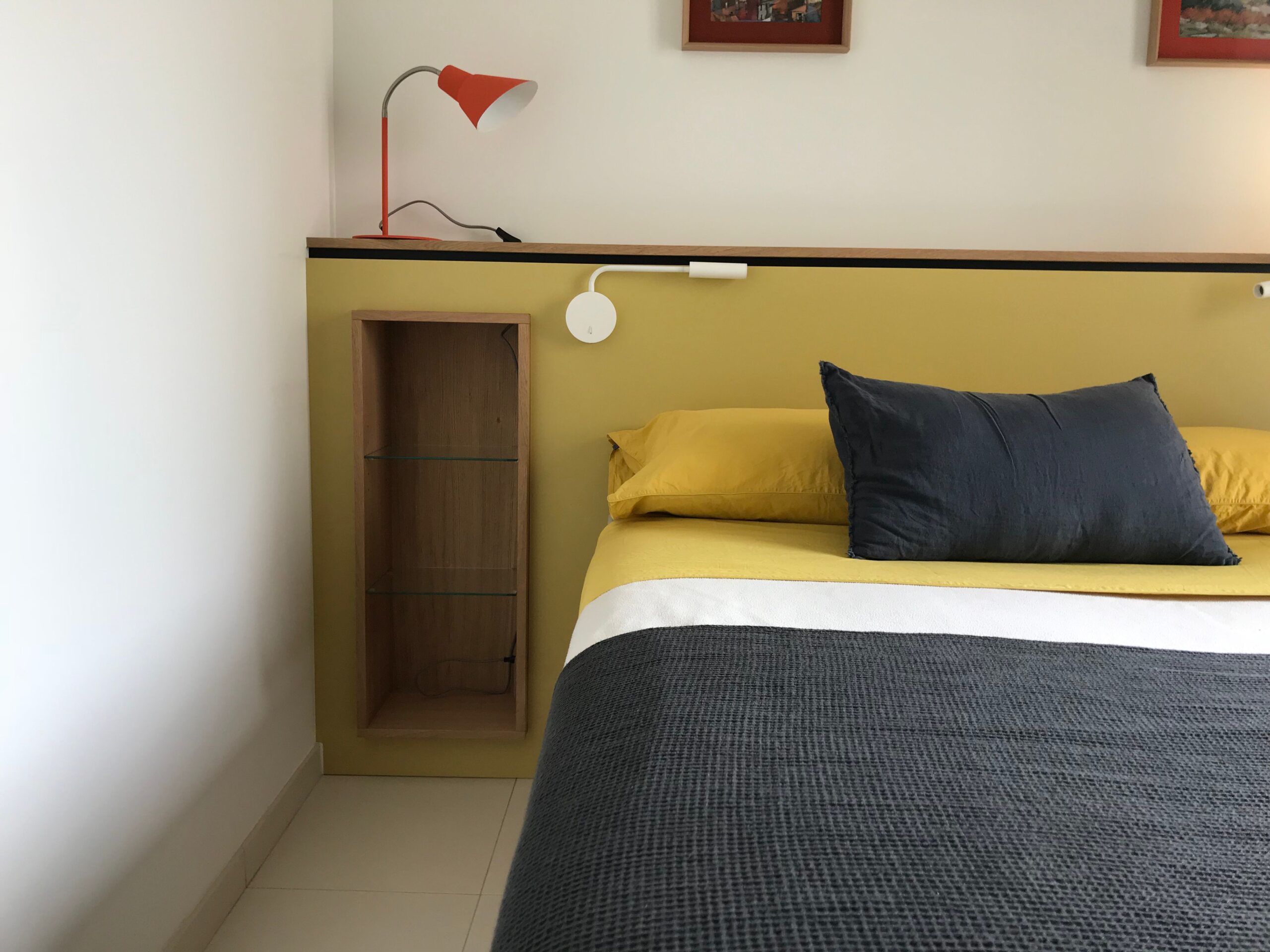 Lit aux draps jaunes et gris, tête de lit moutarde, niche en bois et lampe de bureau rouge.