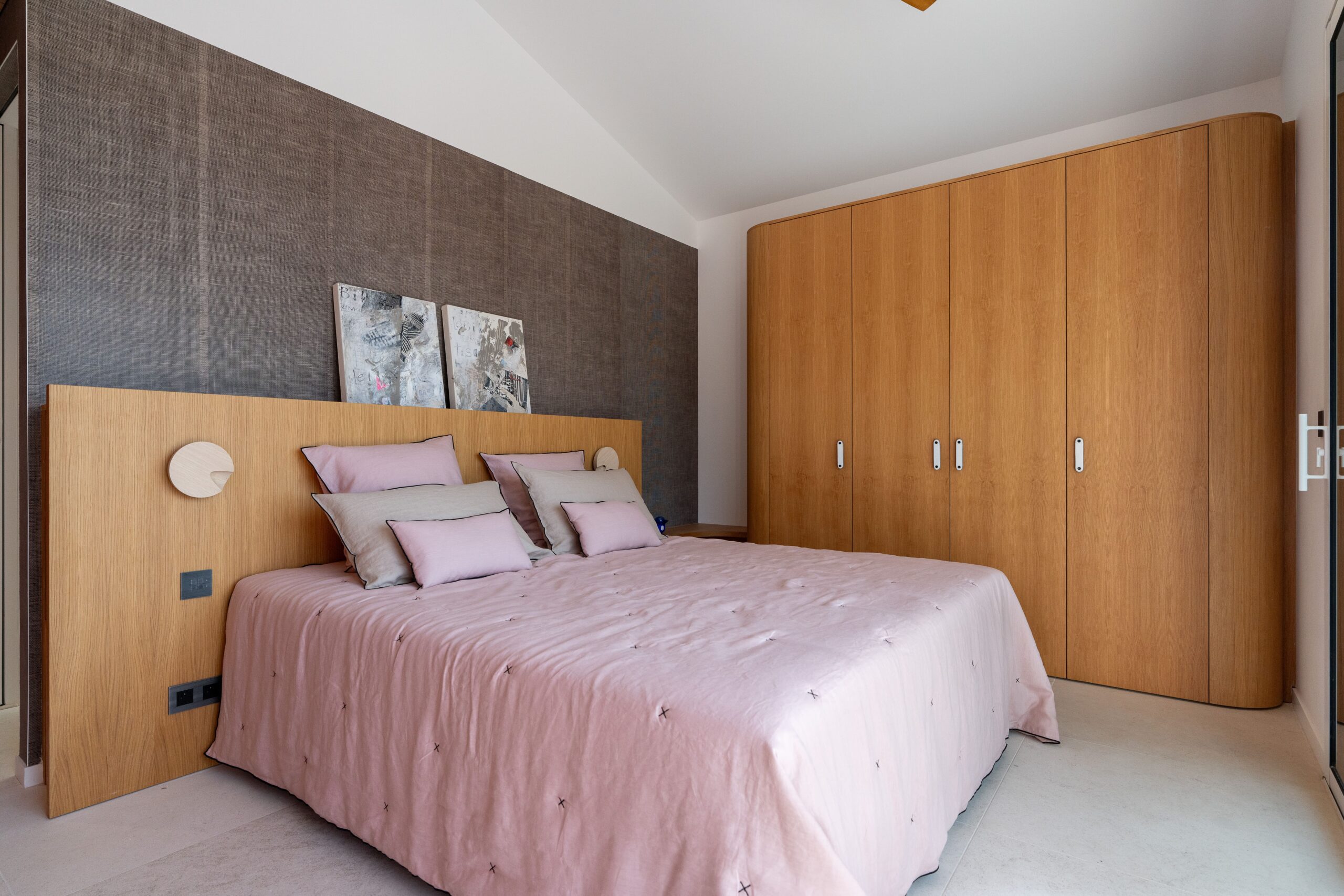 Chambre moderne : lit rose, tête de lit et placard en bois clair sur fond de papier peint gris texturé.