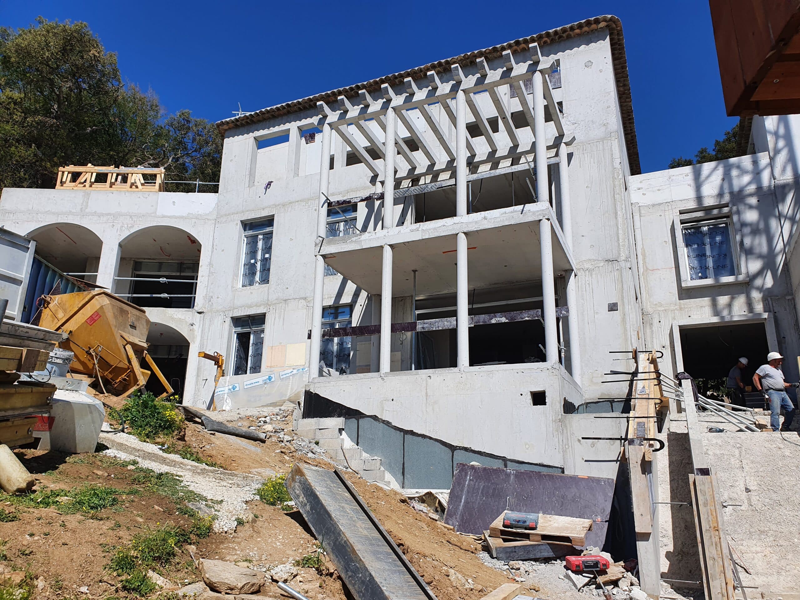 Chantier d'une villa de luxe en béton avec pergola et ouvriers sous un ciel bleu dégagé.