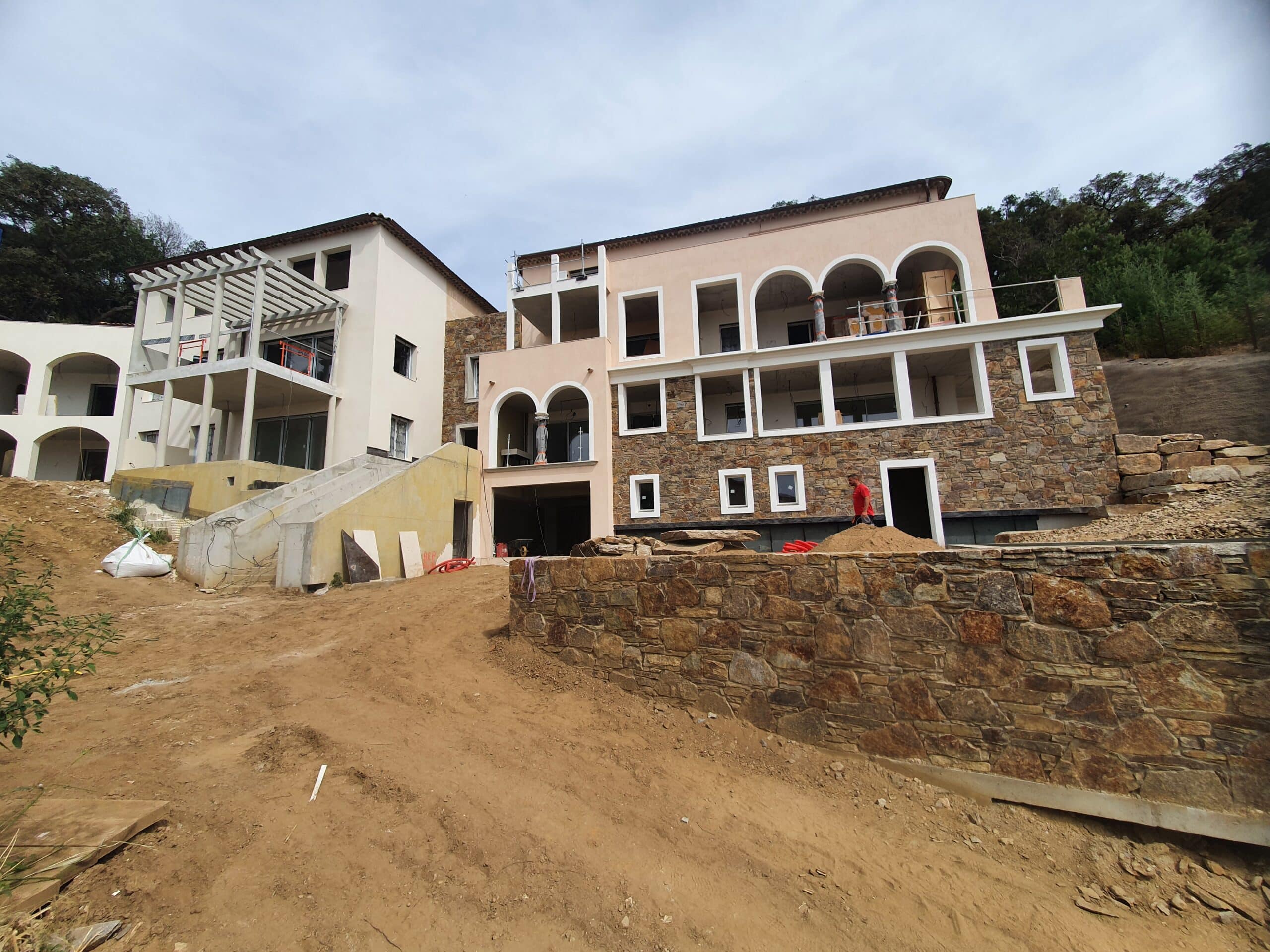 Villa de prestige en construction mêlant pierre et enduit clair sur un terrain escarpé en chantier.