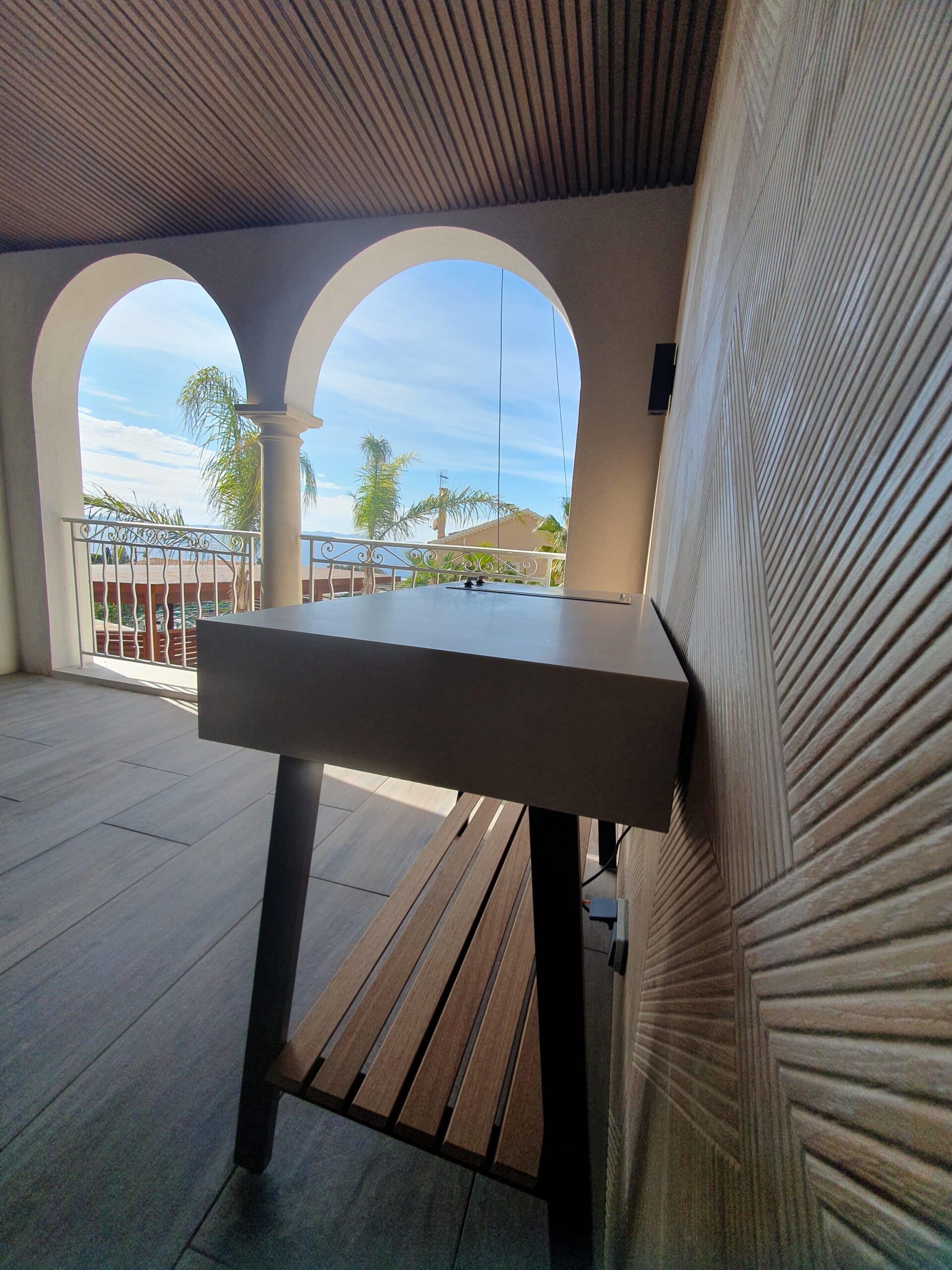 Table de cuisine extérieure sur balcon à arches offrant une vue panoramique sur la mer et les palmiers.