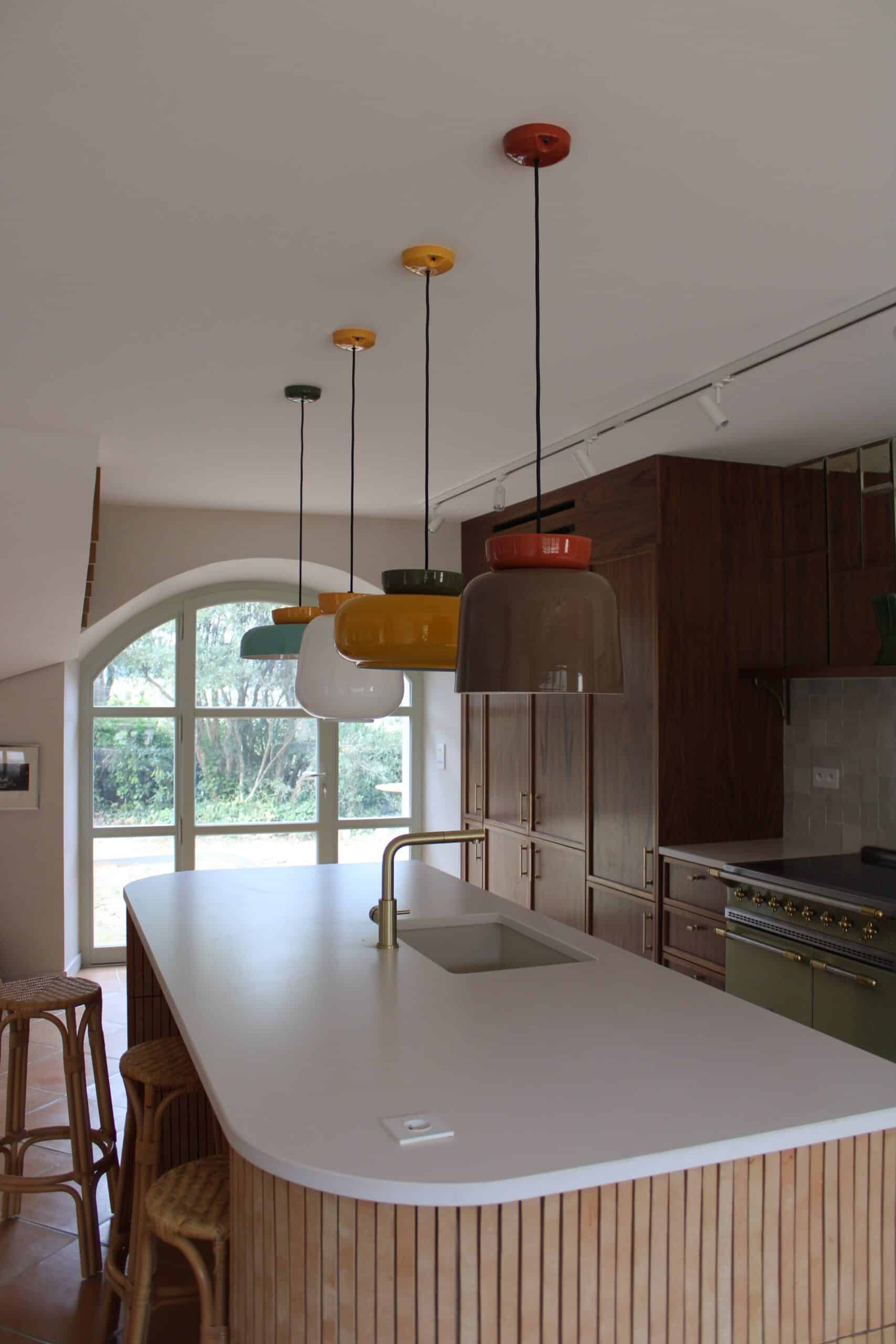 Îlot central blanc, suspensions colorées vintage et meubles bois dans une cuisine lumineuse.