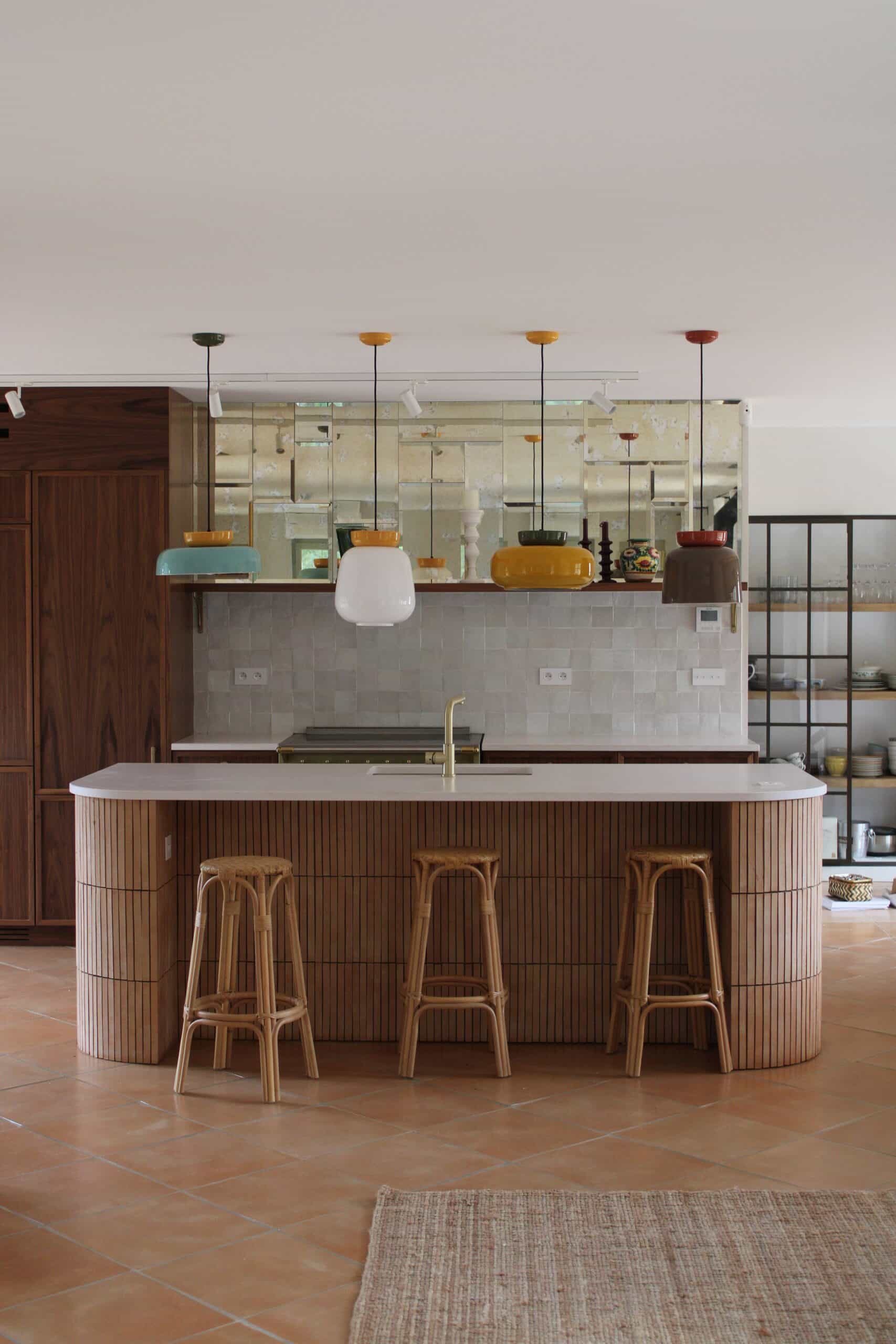 Cuisine moderne avec îlot en bois cannelé, tabourets en rotin, suspensions colorées et sol en terre cuite.