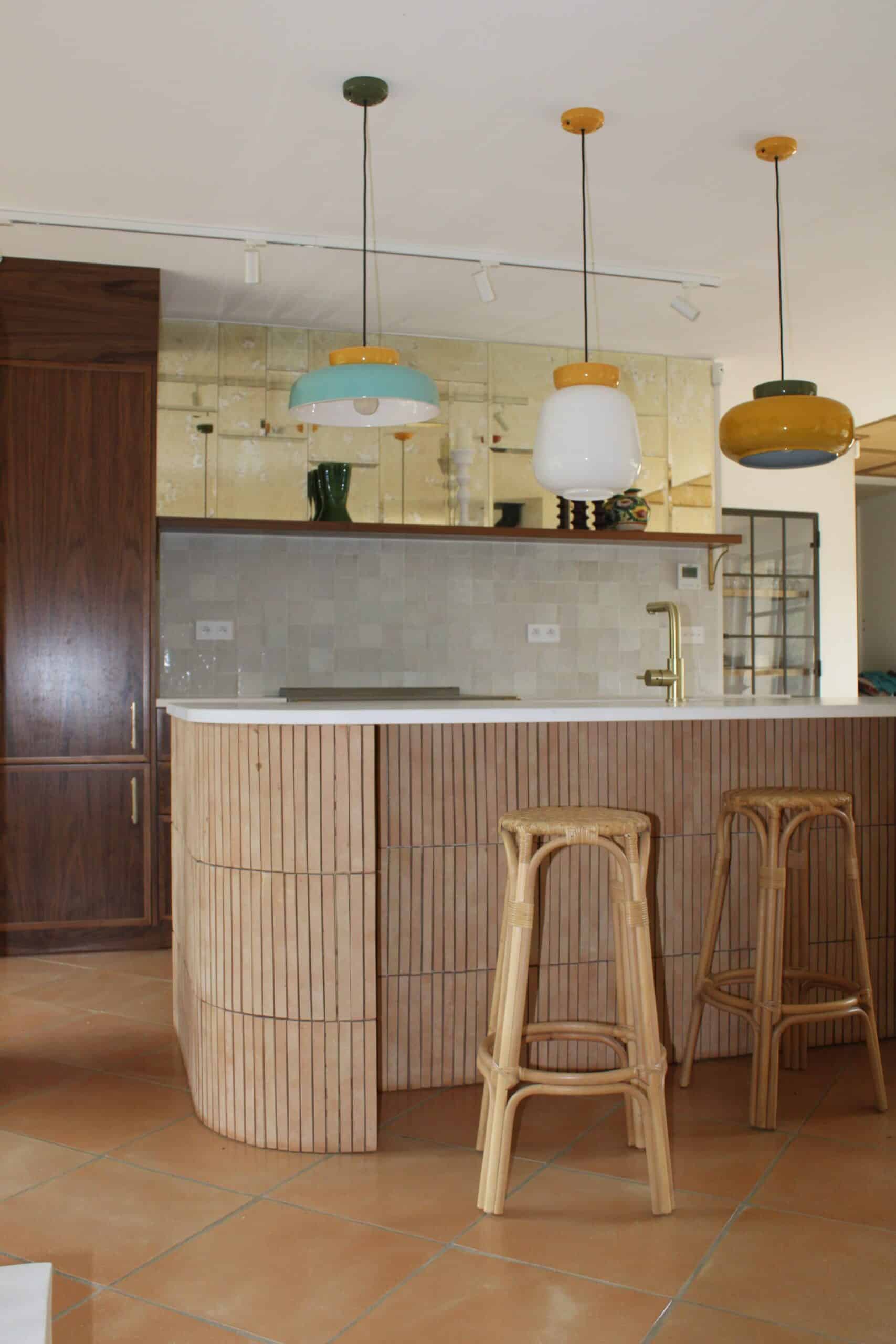 Îlot de cuisine en bois latté, suspensions colorées et tabourets en rotin devant un mur de miroirs vieillis.