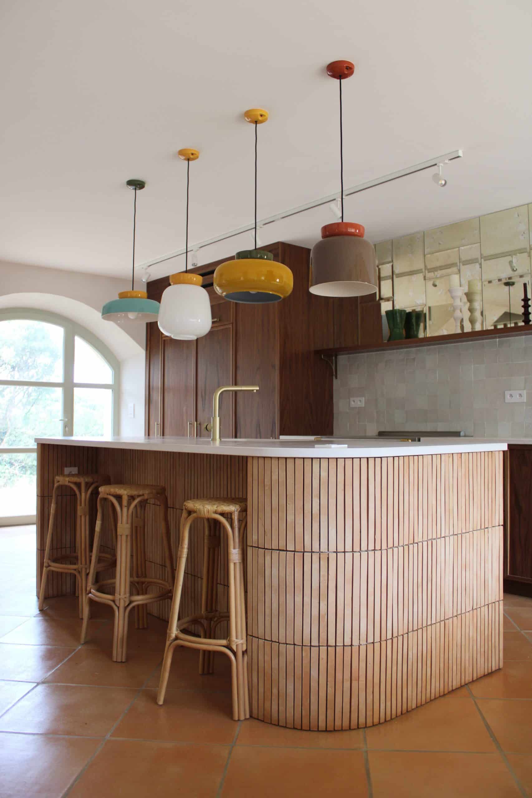 Cuisine moderne avec îlot en lattes de bois, suspensions colorées et tabourets en rotin sur sol en terre cuite.