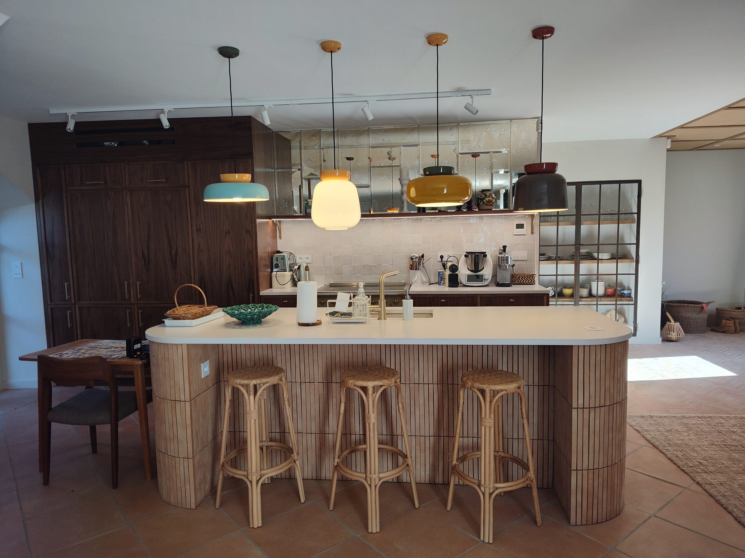 Cuisine moderne avec îlot en bois clair, tabourets en rotin et suspensions colorées design sur sol en terre cuite.