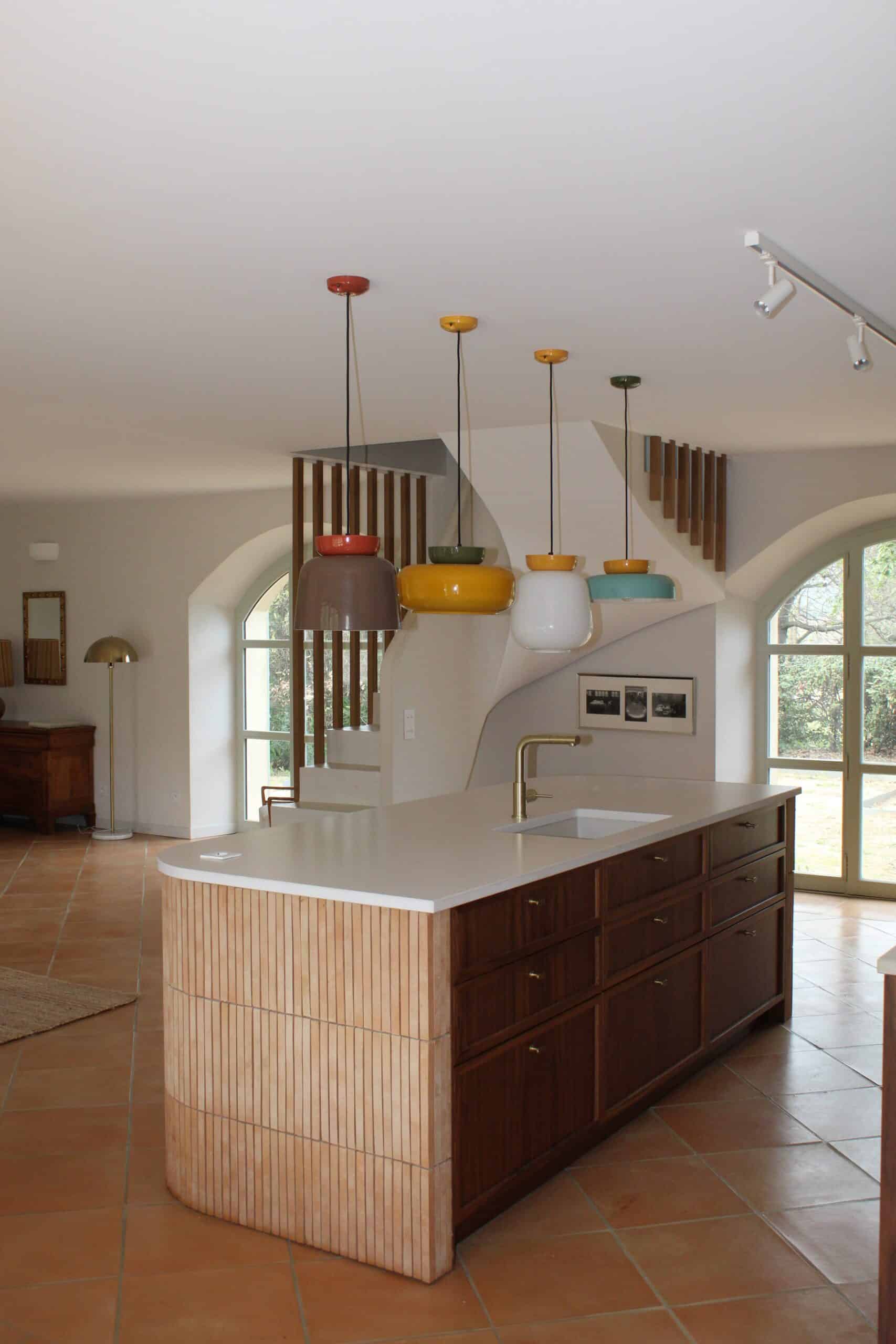 Cuisine moderne avec îlot central en bois, suspensions colorées et sol en terre cuite.