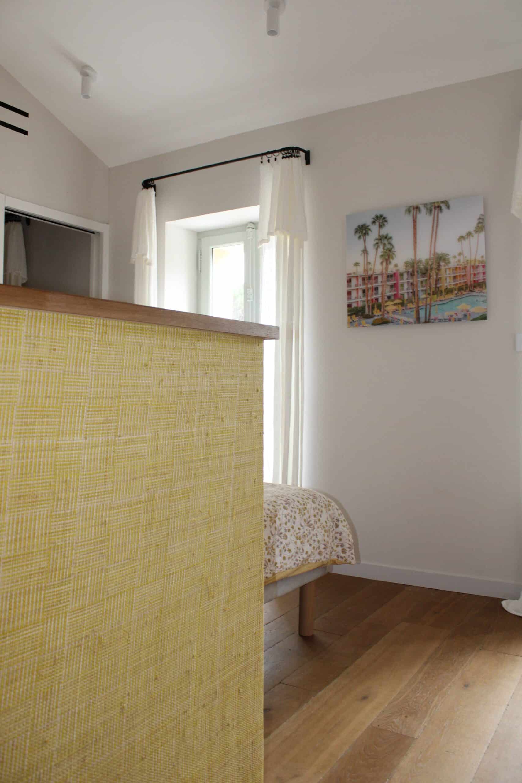 Chambre lumineuse avec muret jaune tressé, lit à fleurs et tableau de palmiers sur mur blanc.