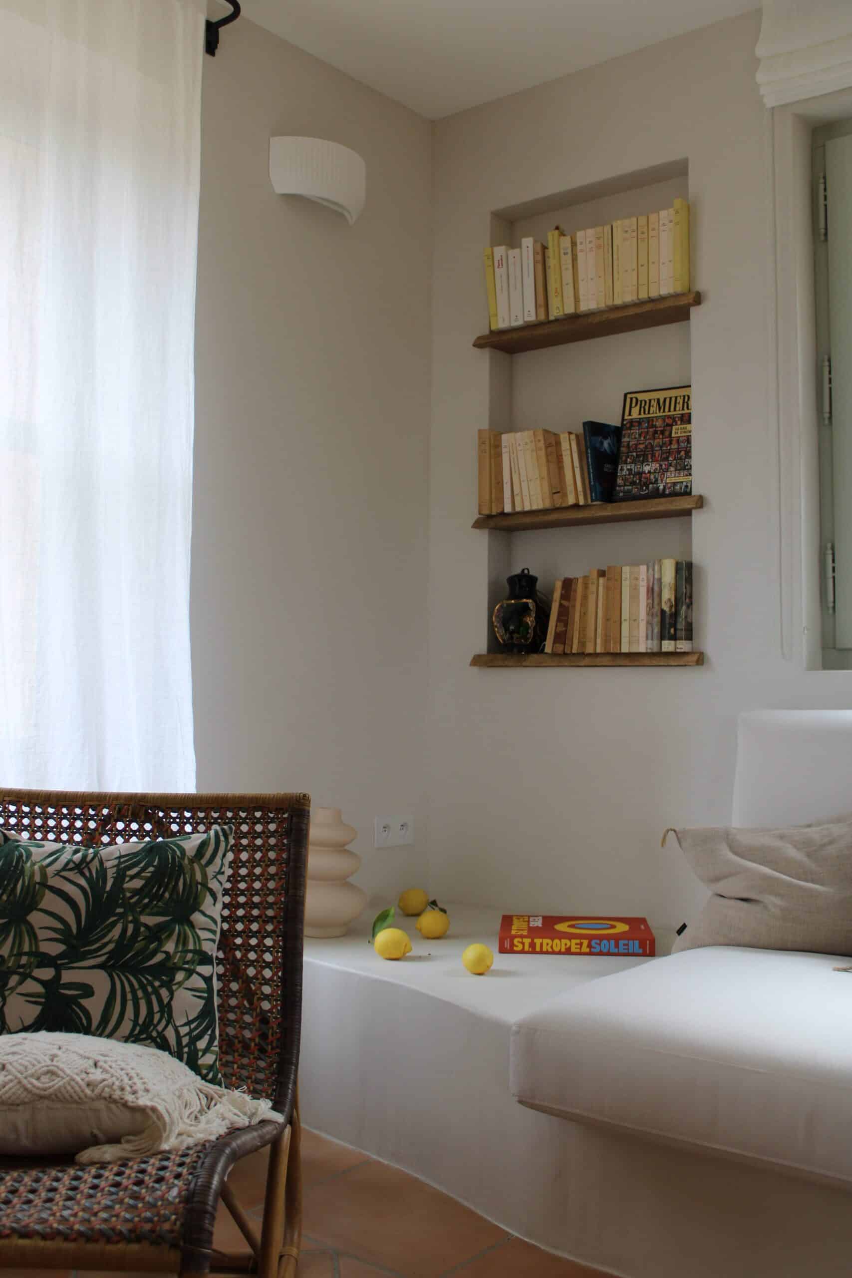 Coin salon serein avec étagères bois, livre ST. TROPEZ SOLEIL, citrons et fauteuil rotin motif palmier.
