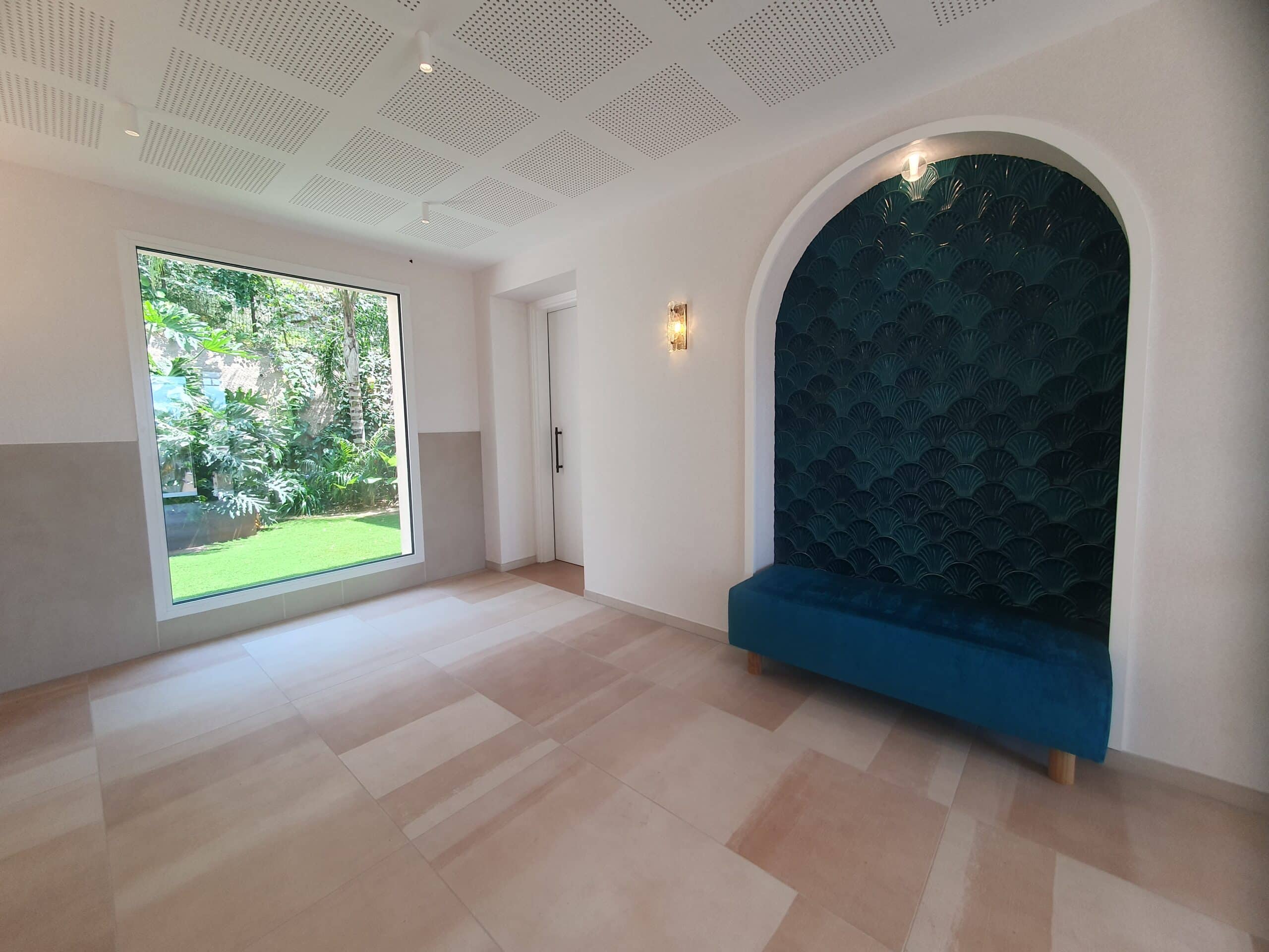 Intérieur moderne avec alcôve à écailles bleues, banquette en velours et grande fenêtre ouvrant sur un jardin tropical.