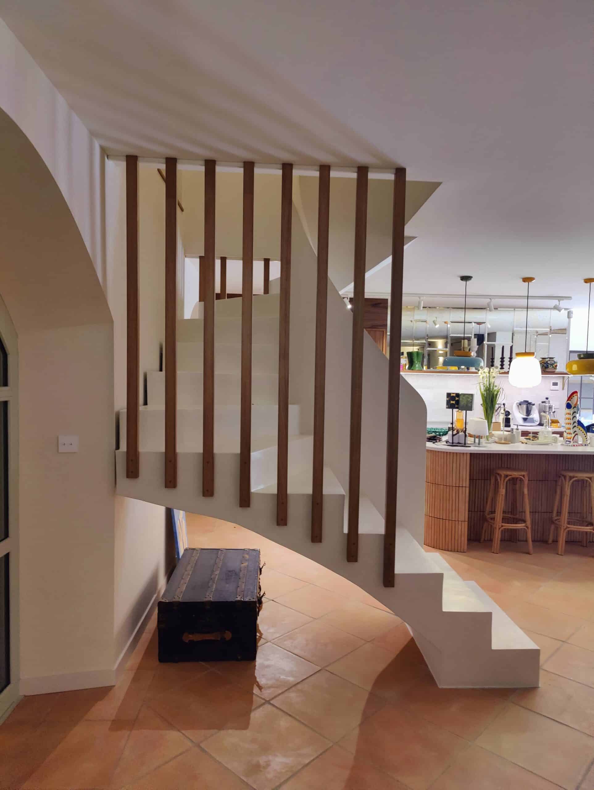 Escalier blanc sculptural à lattes de bois, coffre vintage et cuisine ouverte avec îlot central en bois.