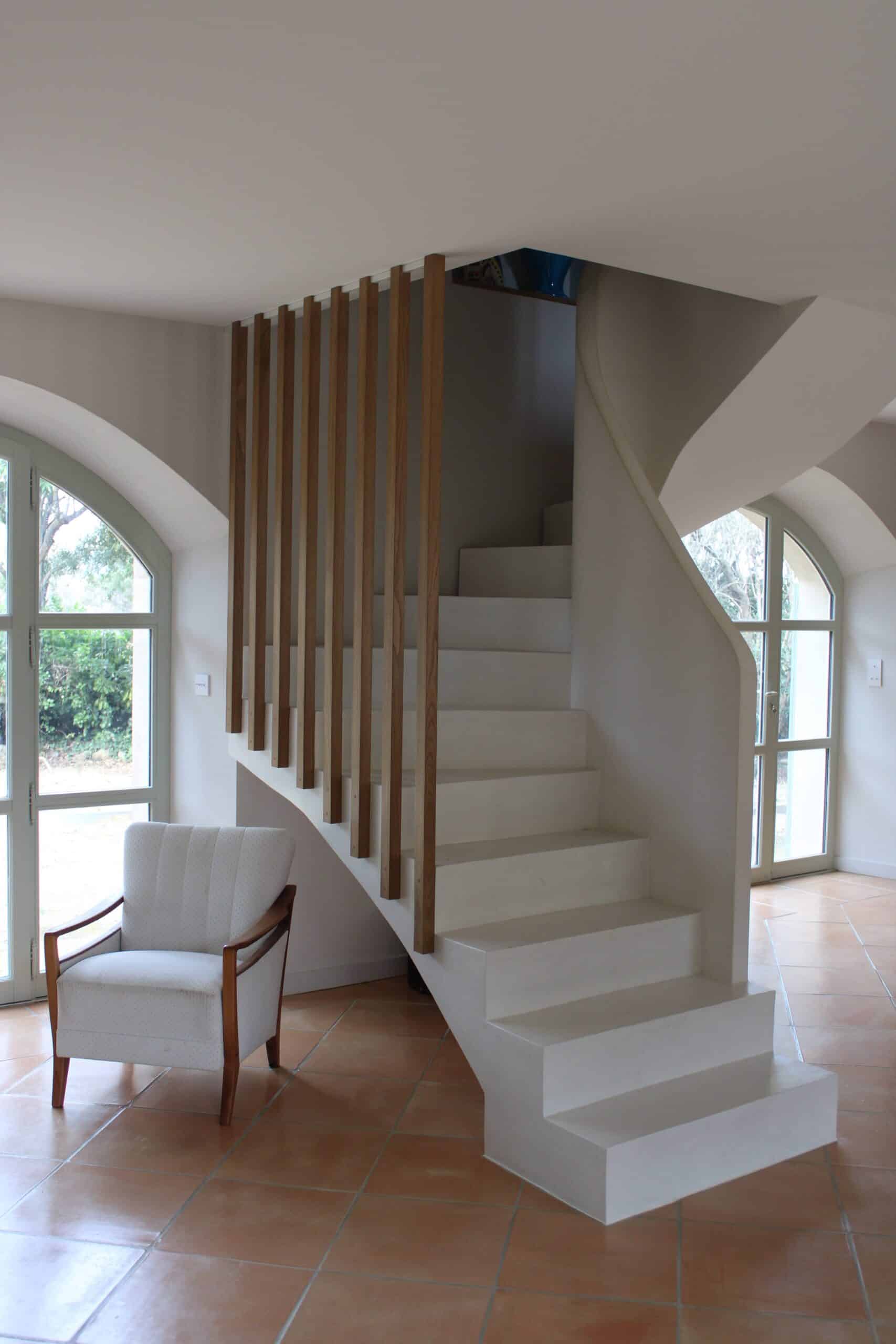 Escalier blanc moderne avec tasseaux bois, fauteuil blanc et sol en terre cuite près de fenêtres cintrées.