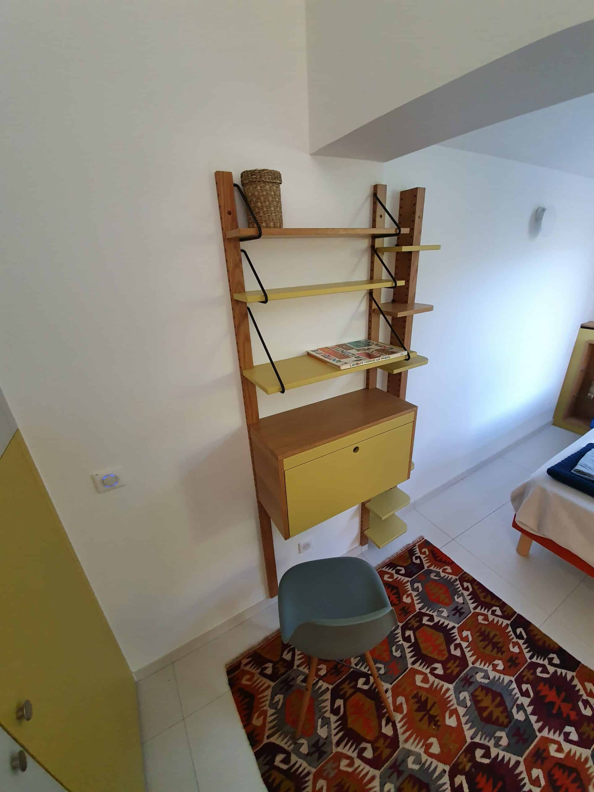 Étagère murale en bois et jaune avec bureau escamotable, chaise grise et tapis coloré à motifs.