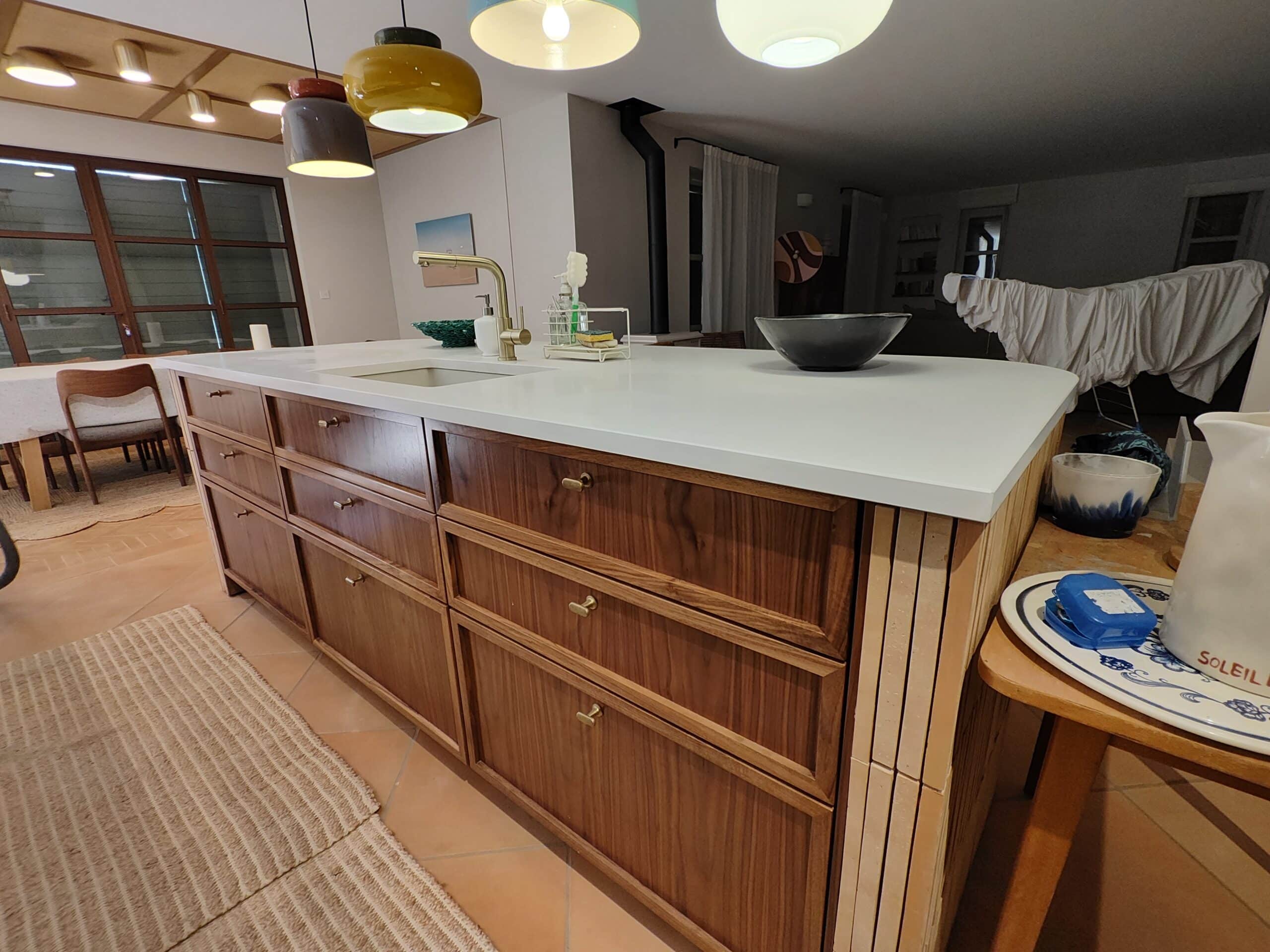 Îlot central en bois foncé, plan de travail blanc et robinet doré sous des suspensions colorées.