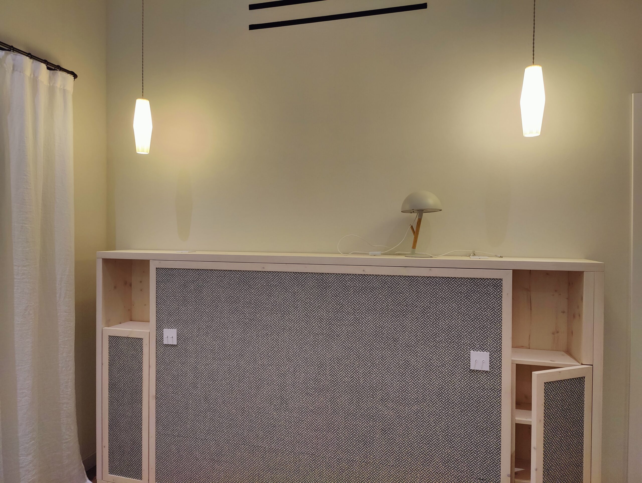 Lit escamotable à motifs gris, étagères bois et luminaires suspendus dans un décor épuré.