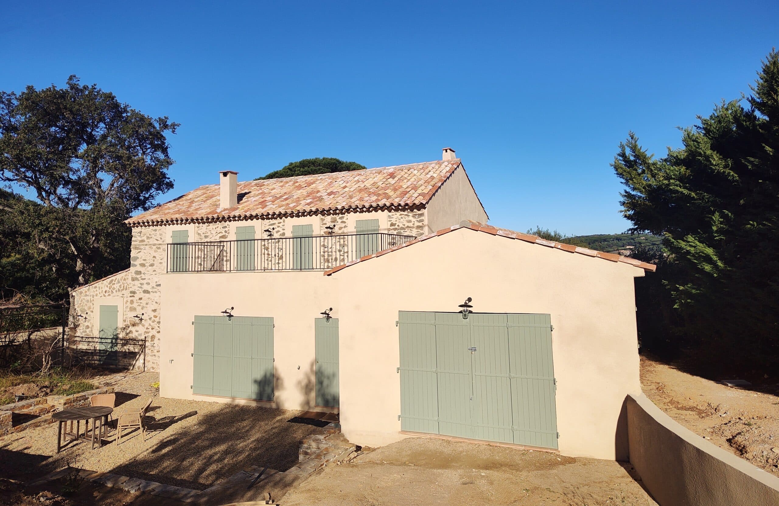 Maison provençale en pierre avec volets vert olive, toit en terre cuite et terrasse sous un ciel bleu.