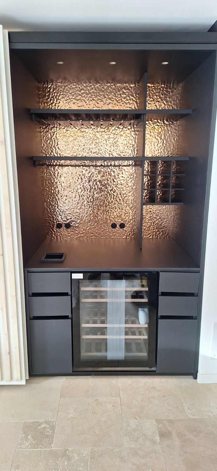 Bar moderne encastré avec crédence en cuivre martelé, porte-verres et cave à vin intégrée noire.