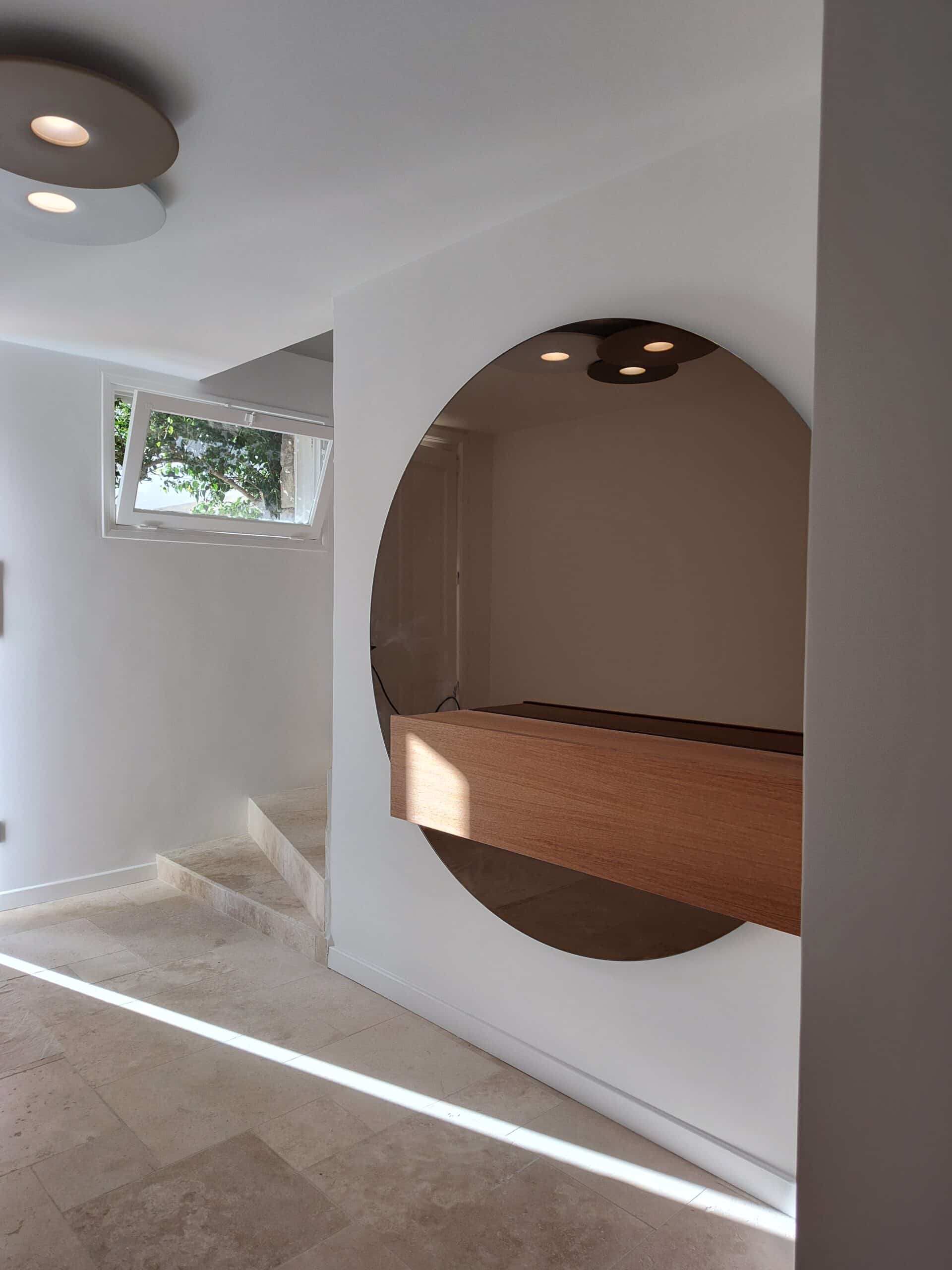 Couloir moderne avec grand miroir circulaire, console en bois suspendue, escalier et sol en pierre travertin.