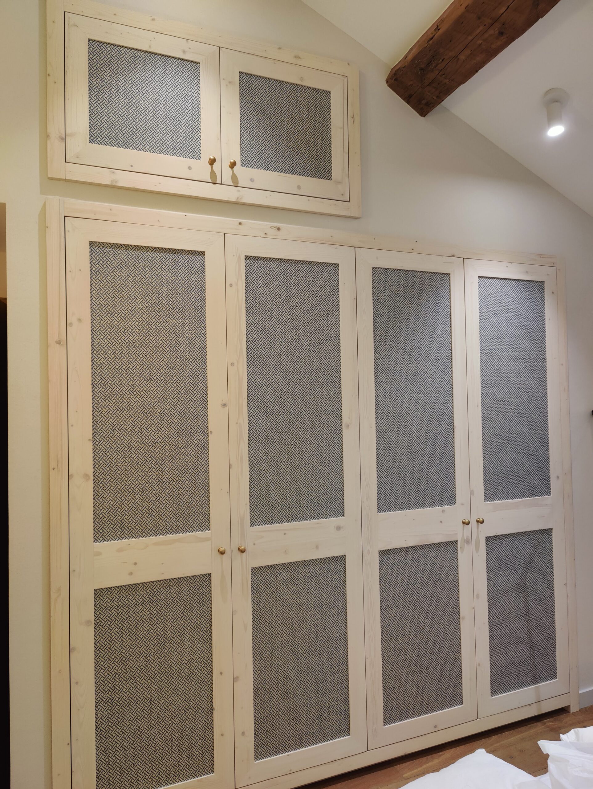 Placard sur mesure en bois clair avec portes grillagées à motif géométrique et boutons dorés sous poutre apparente.