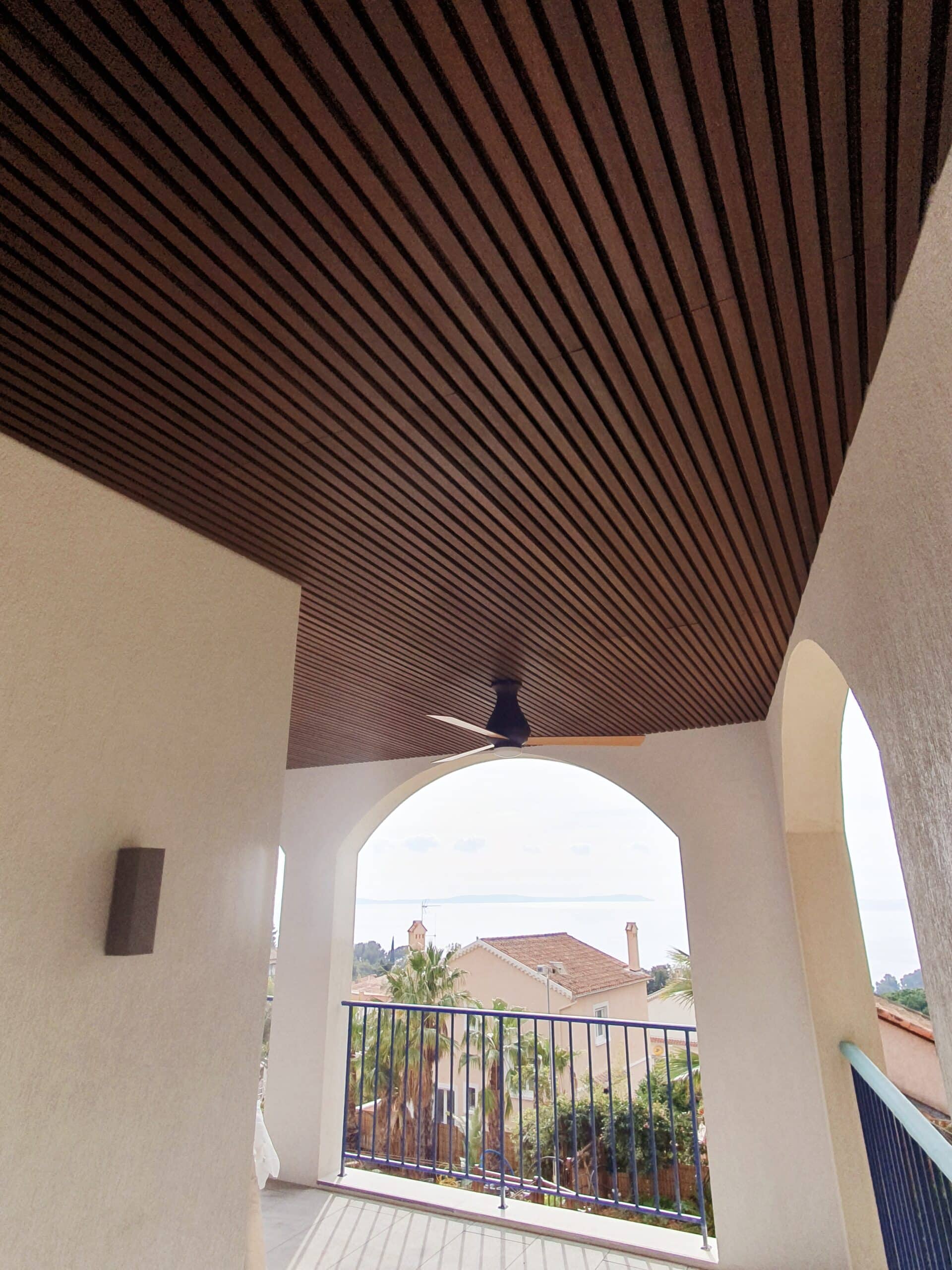 Terrasse moderne avec plafond à claire-voie en bois, ventilateur de plafond et vue mer panoramique via une arche.