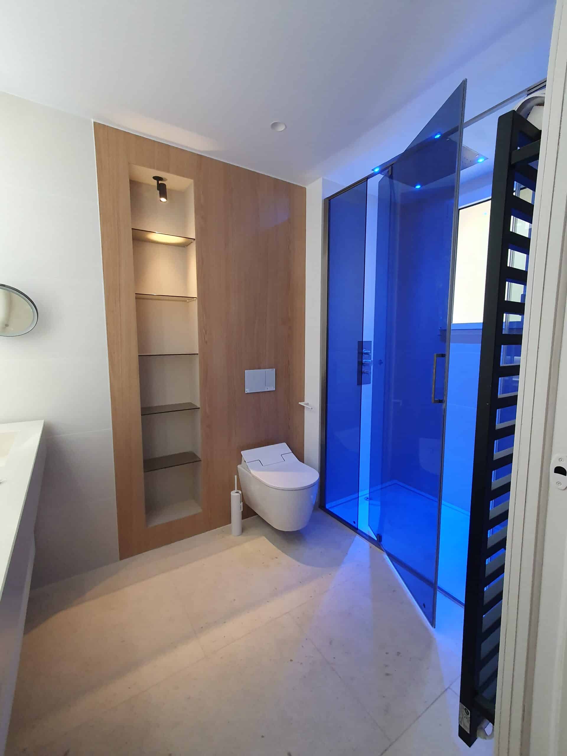 Salle de bain moderne : douche à éclairage bleu, WC suspendu et étagères en bois clair intégrées.