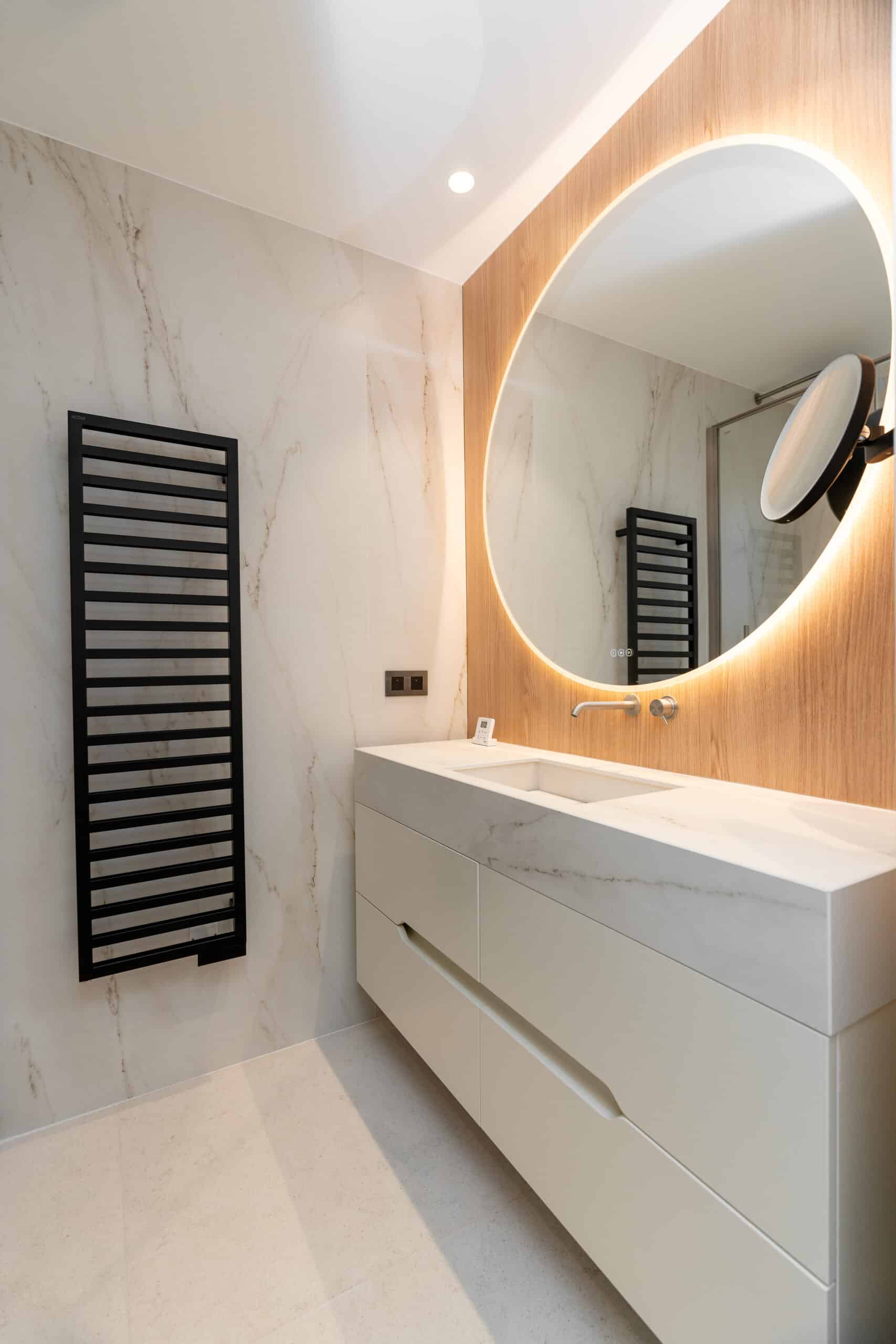 Salle de bain moderne : miroir rétroéclairé rond, meuble vasque marbre blanc et radiateur sèche-serviette noir.