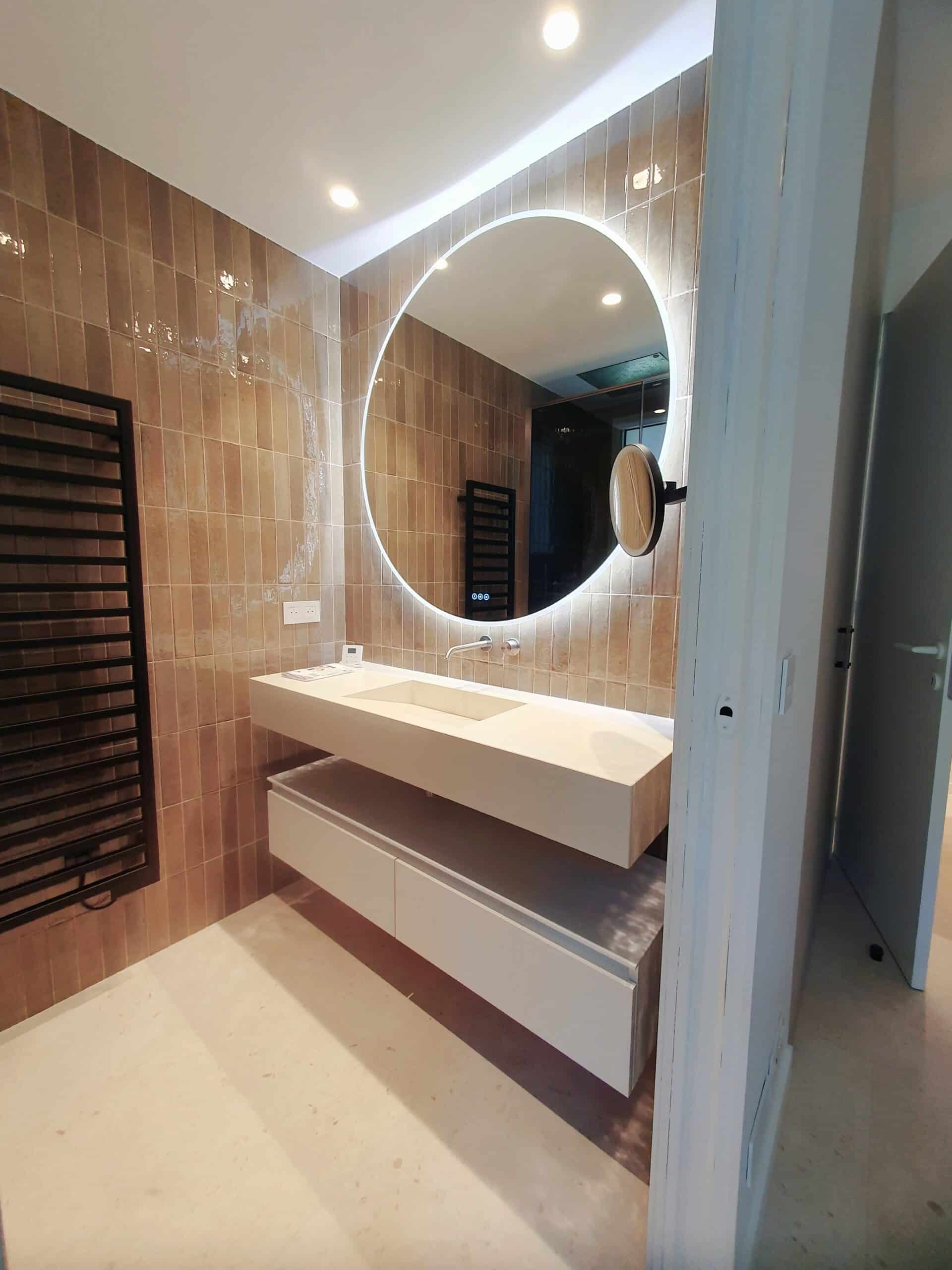 Salle de bain chic avec miroir rond lumineux, vasque blanche suspendue et carrelage mural brun.