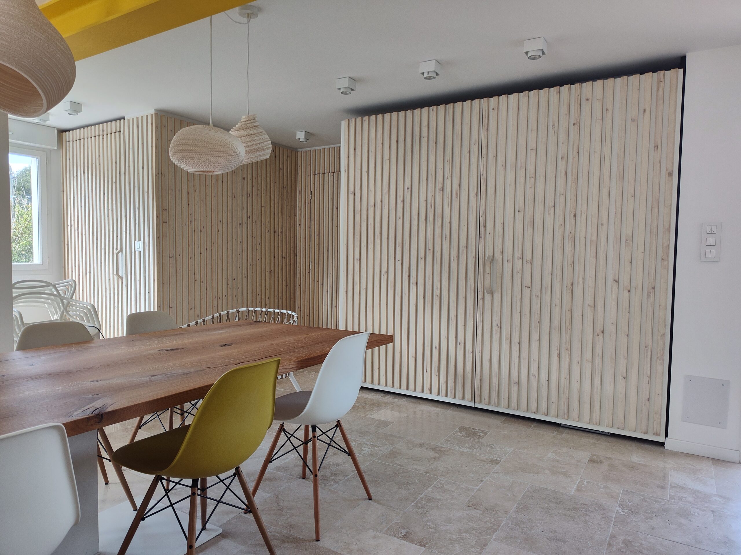 Salle à manger moderne avec table en bois massif, chaises design et mur décoratif en tasseaux de bois clair.