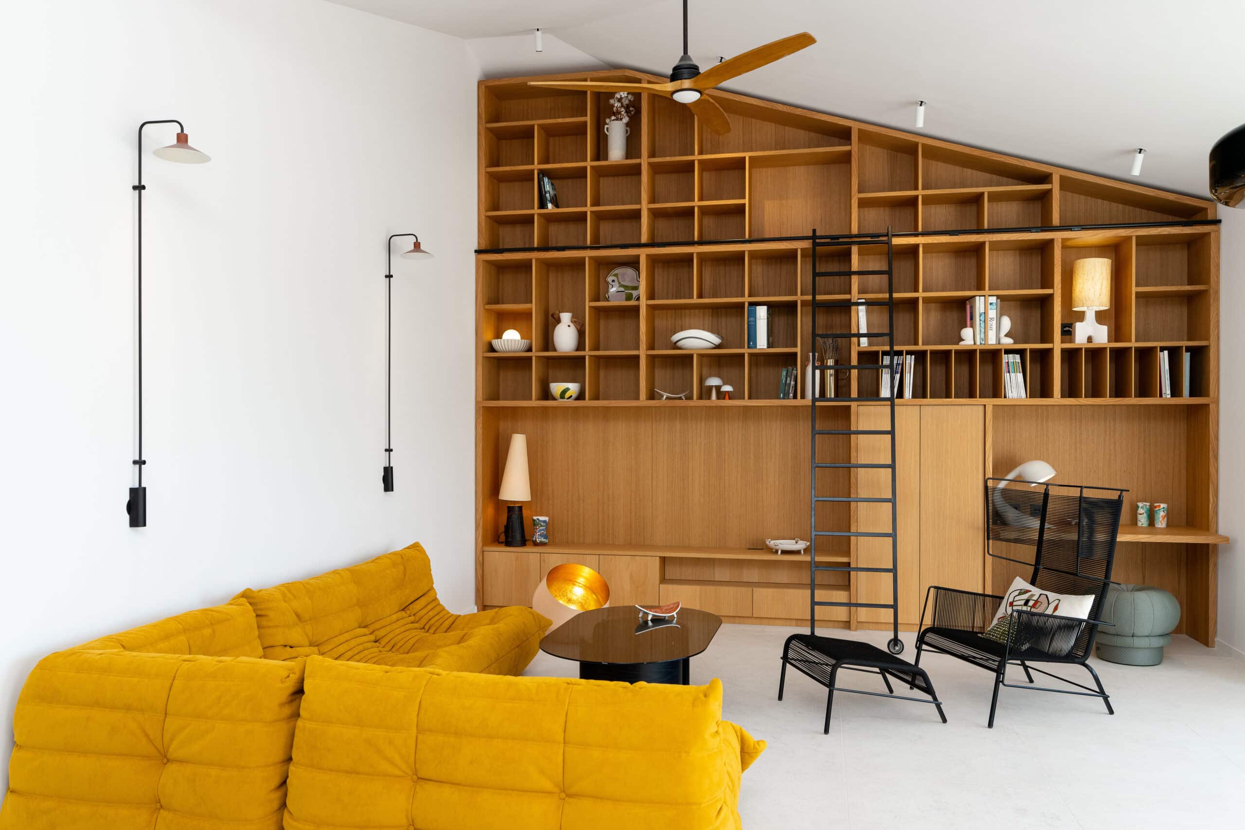 Salon moderne avec canapé jaune capitonné, grande bibliothèque en bois sur mesure et échelle noire coulissante.