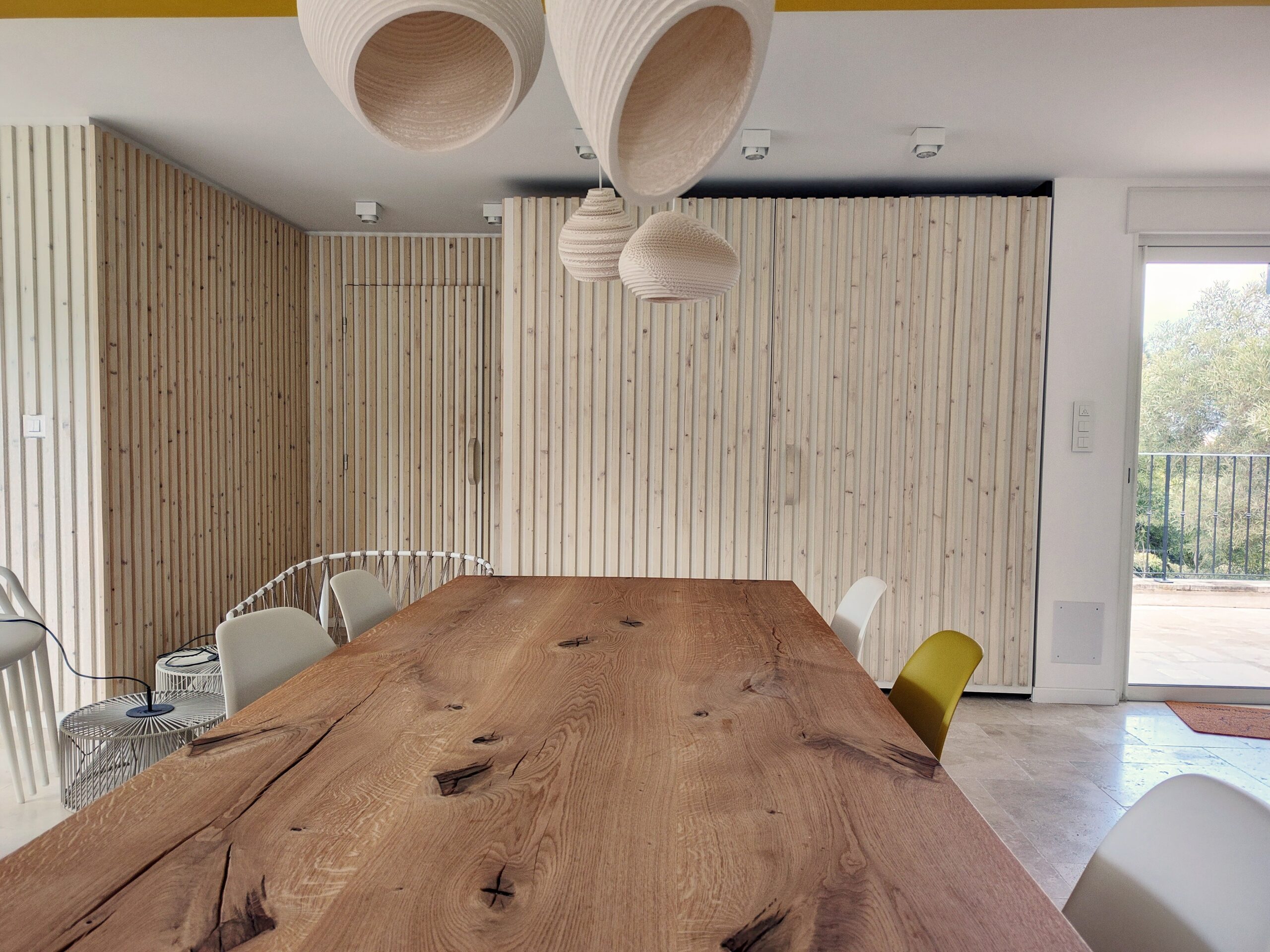 Table en bois massif, chaises design et luminaires suspendus devant un mur en tasseaux de bois clair.
