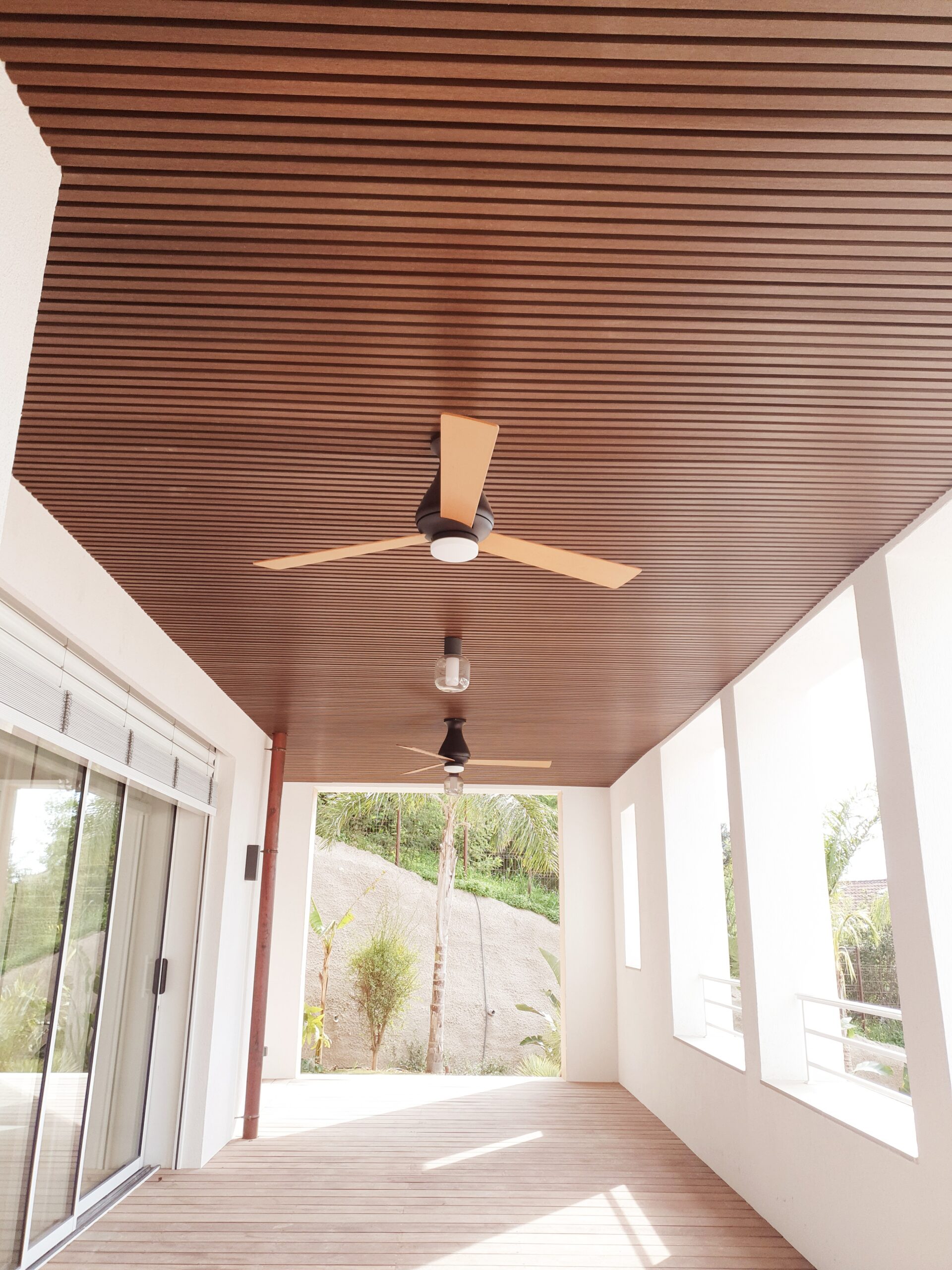 Terrasse moderne avec plafond à lattes de bois, ventilateurs de plafond et vue sur un jardin vallonné.