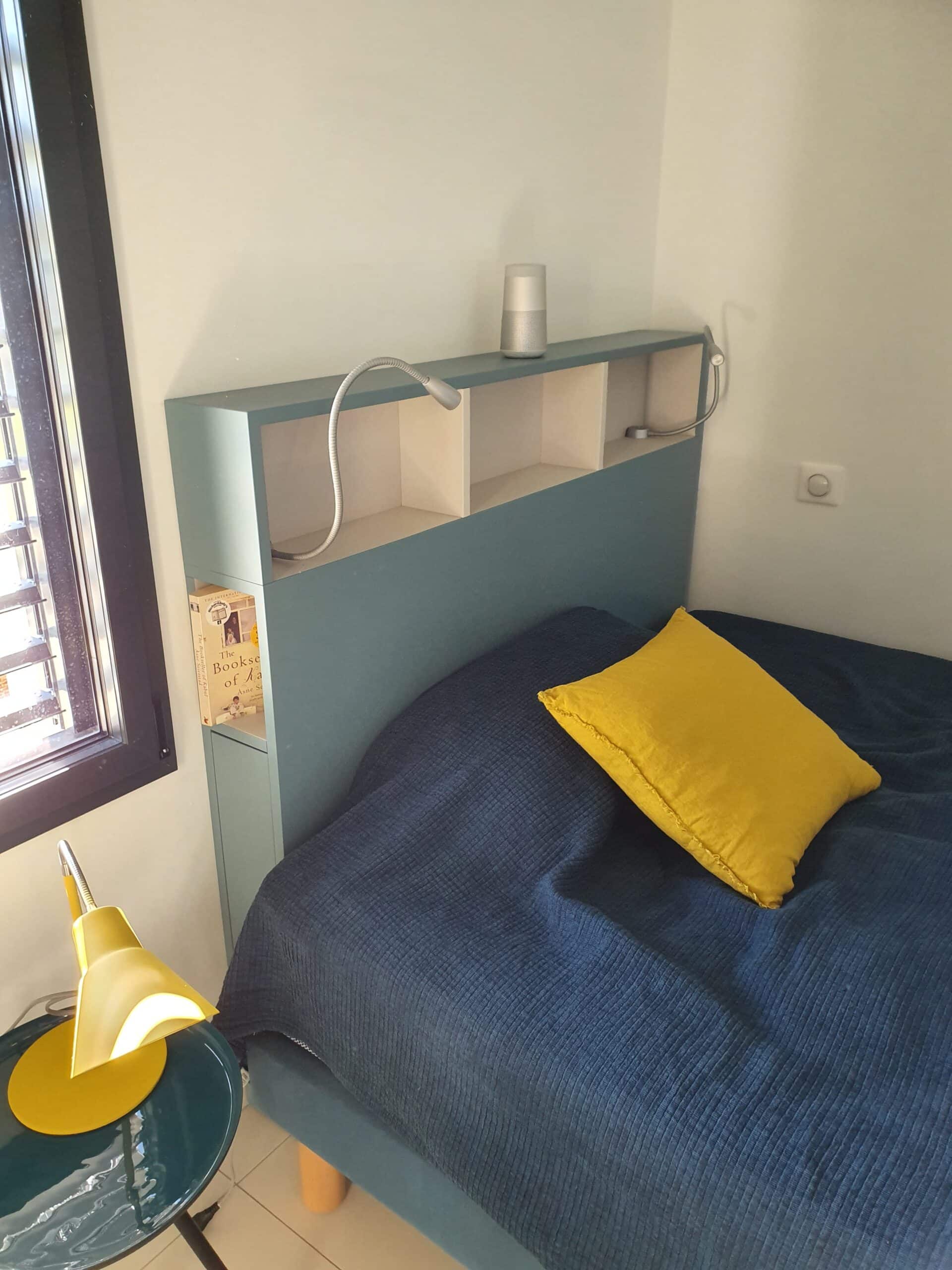 Lit bleu, coussin jaune et tête de lit bleue avec étagères et lampes de lecture intégrées.