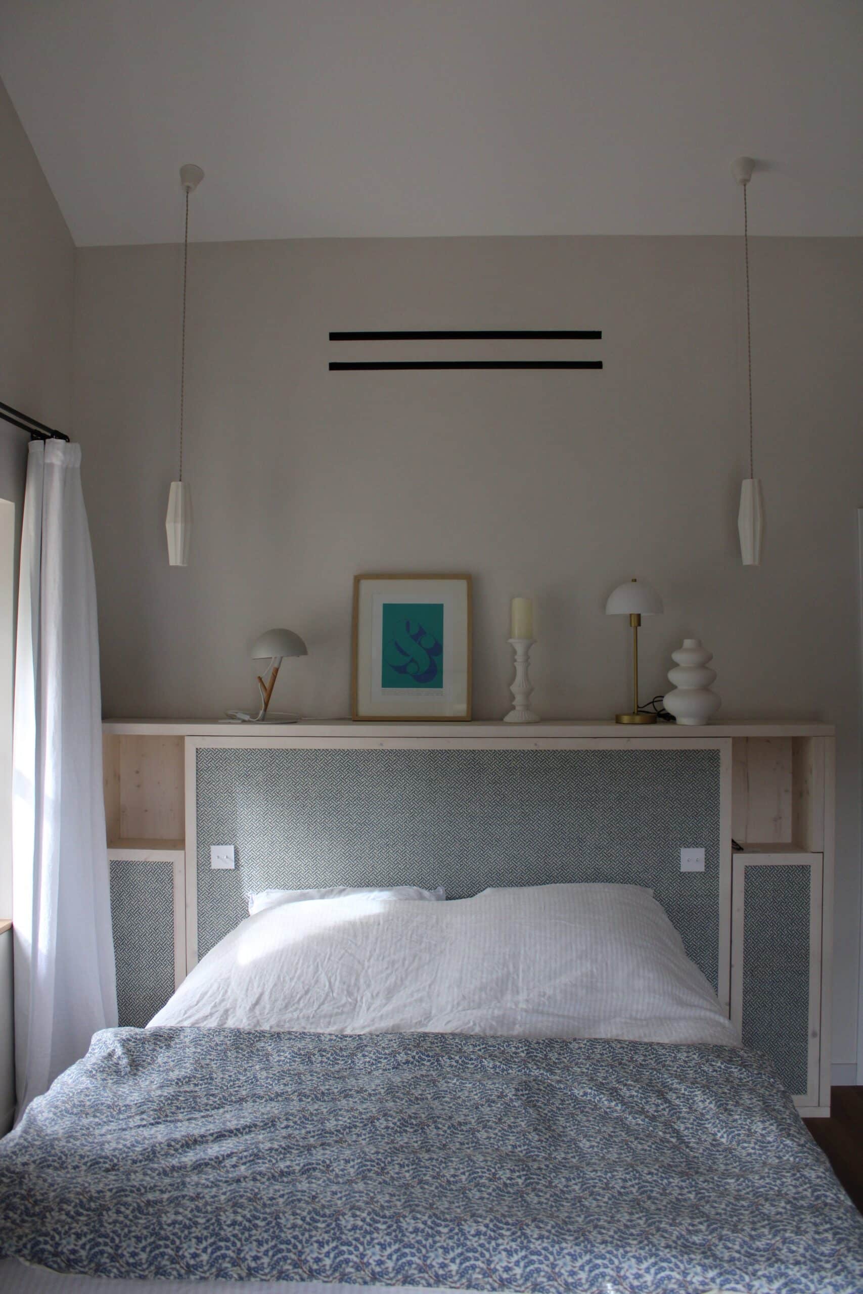 Lit avec tête de lit en bois et tissu bleu, linge de lit fleuri et suspensions blanches dans une chambre épurée.