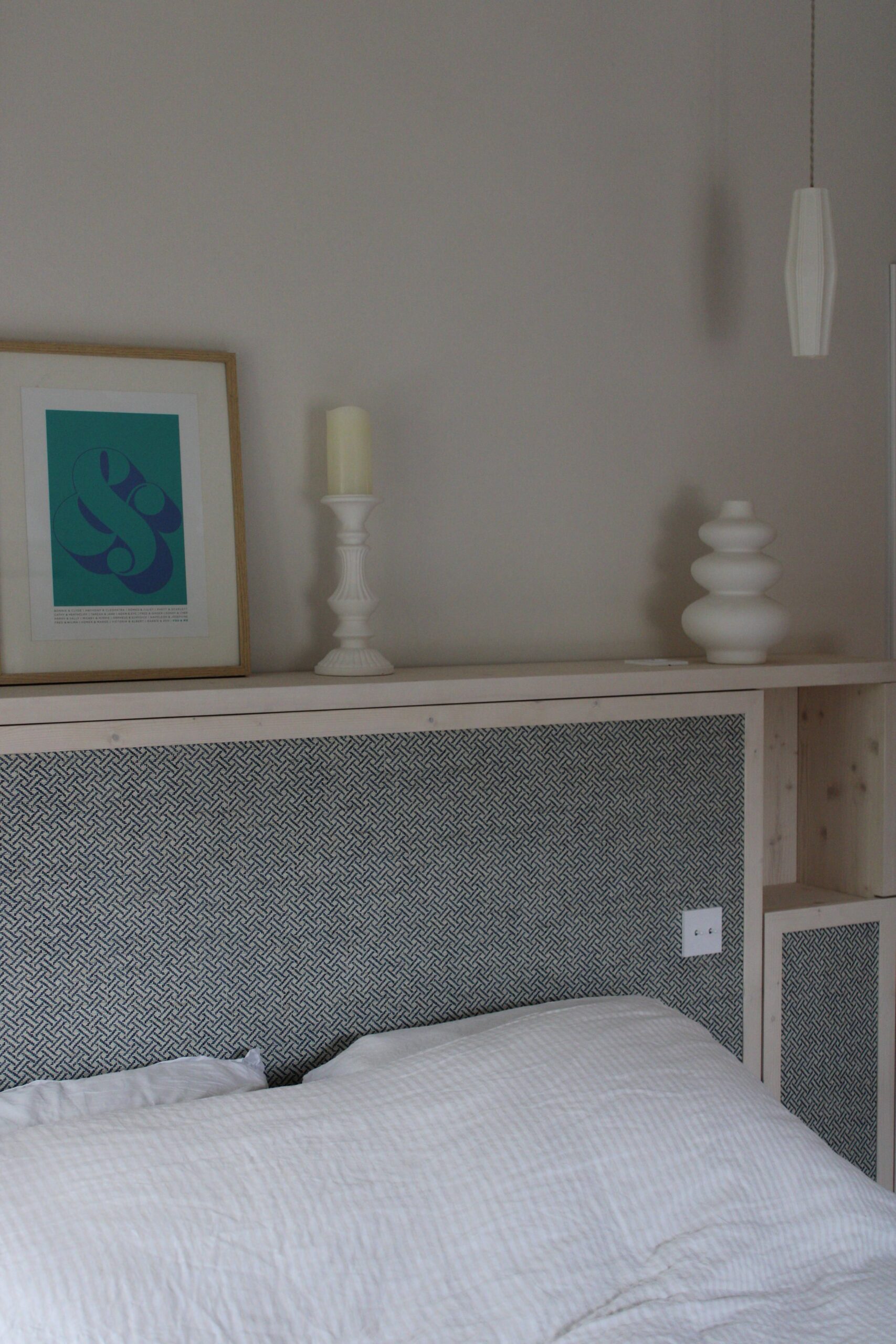 Tête de lit bois, tissu bleu à motifs, étagère avec cadre graphique et objets déco blancs minimalistes.