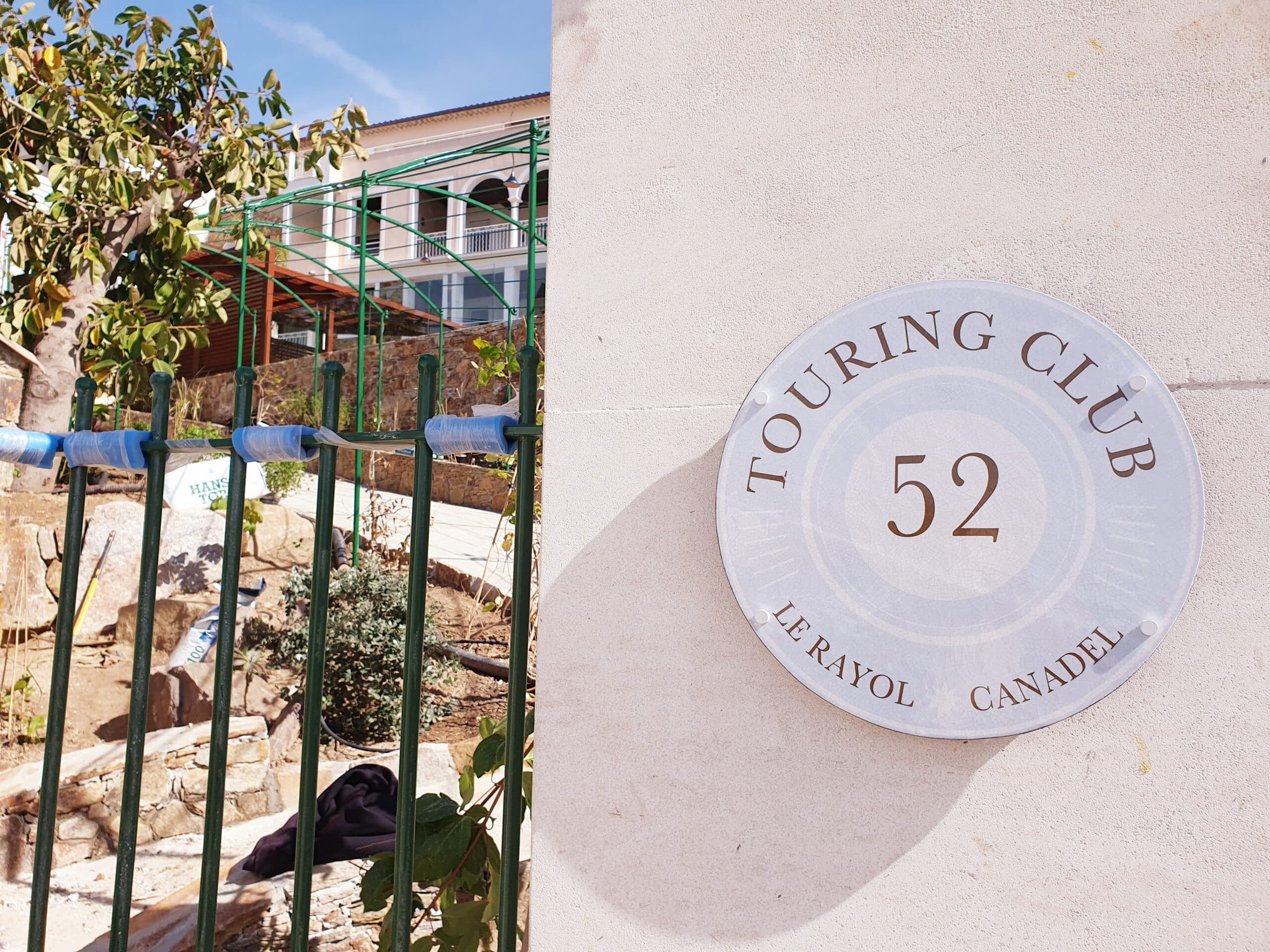 Plaque ronde Touring Club 52, Le Rayol Canadel, fixée sur un mur clair à côté d'une clôture verte.